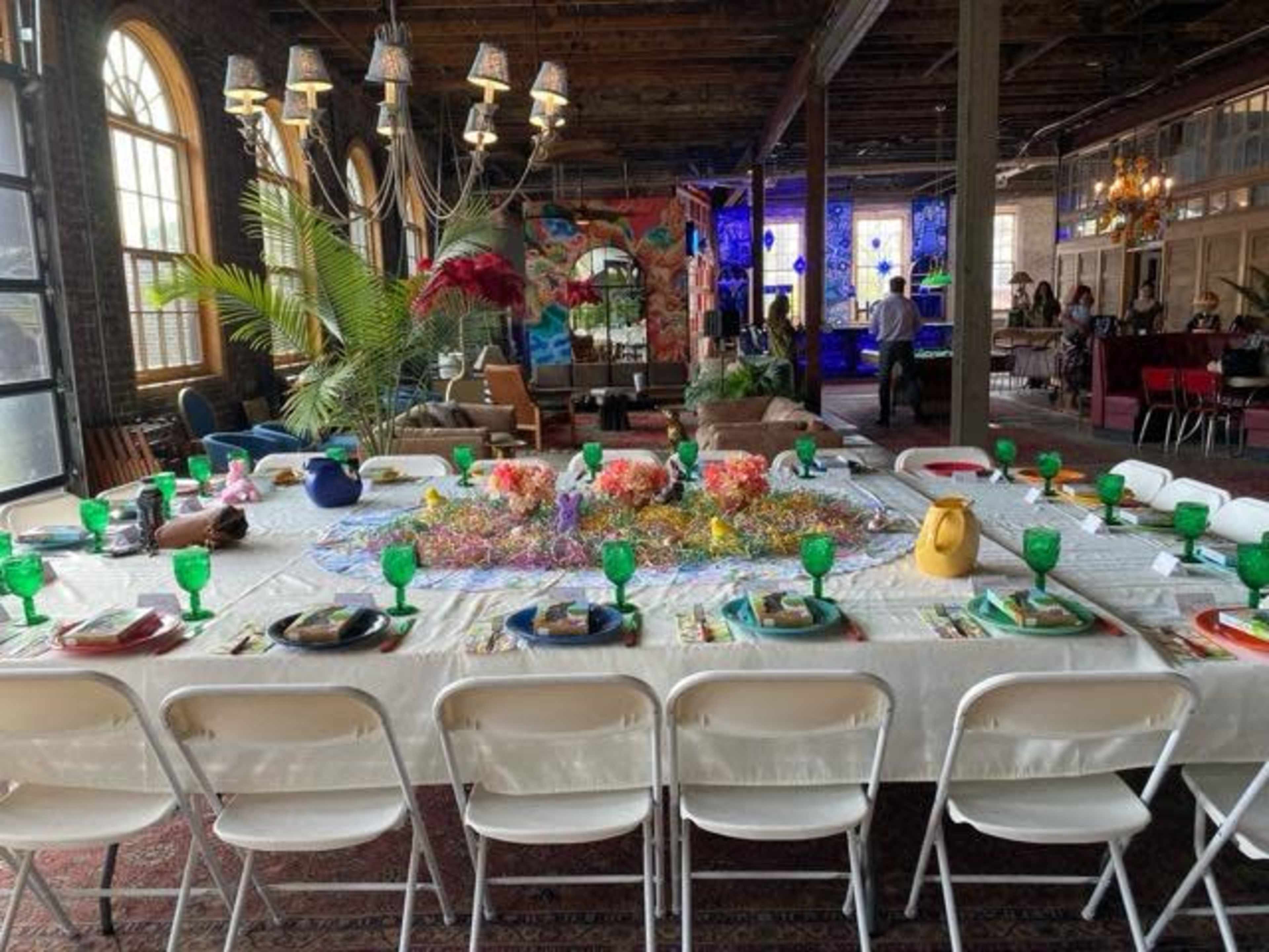 A long dining table is set with green glassware and colorful decorations in a spacious room with large windows and various lighting fixtures.