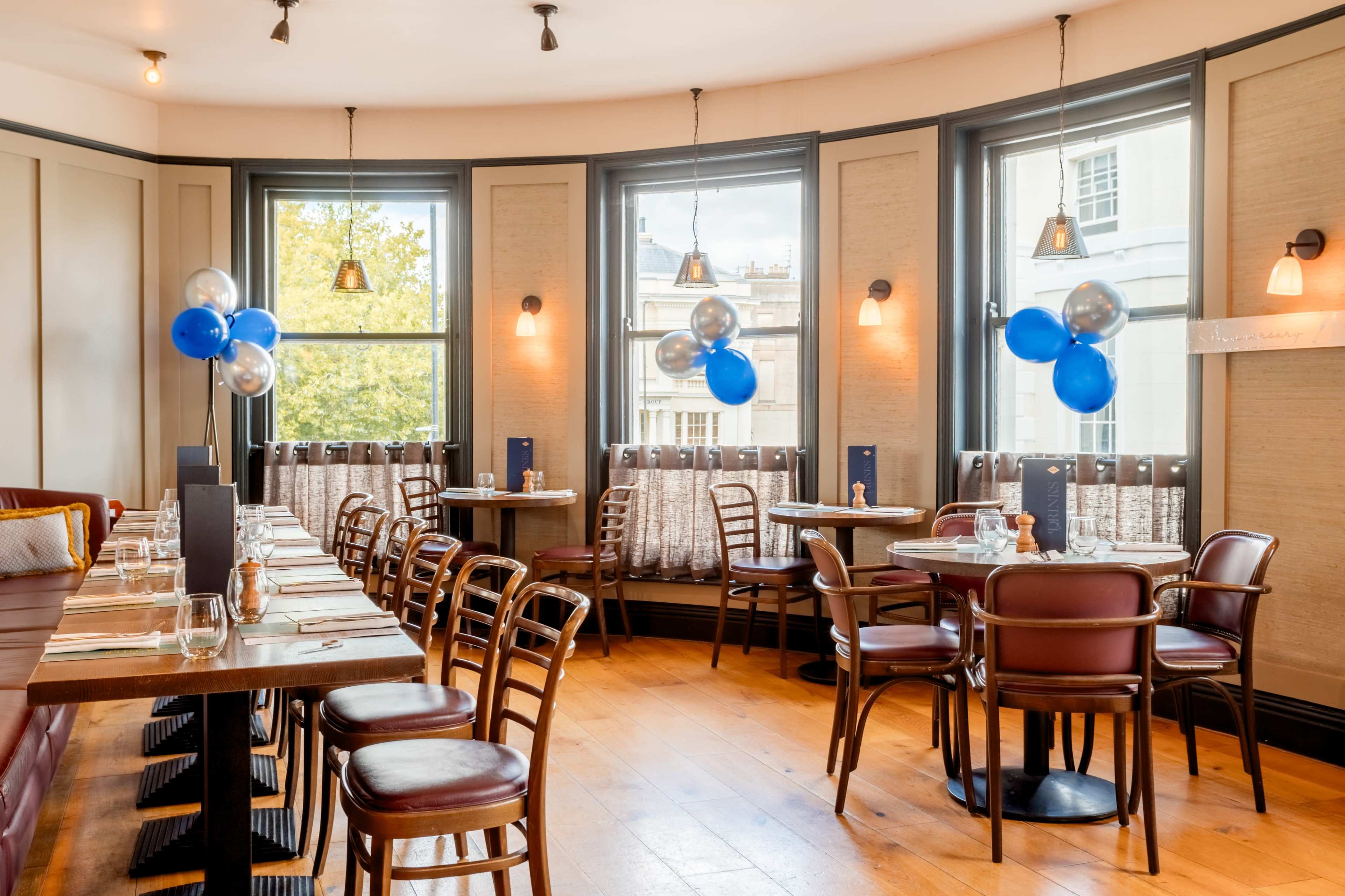 The image depicts a restaurant interior with wooden tables and chairs arranged in a bright corner, featuring decorations of blue and silver balloons.