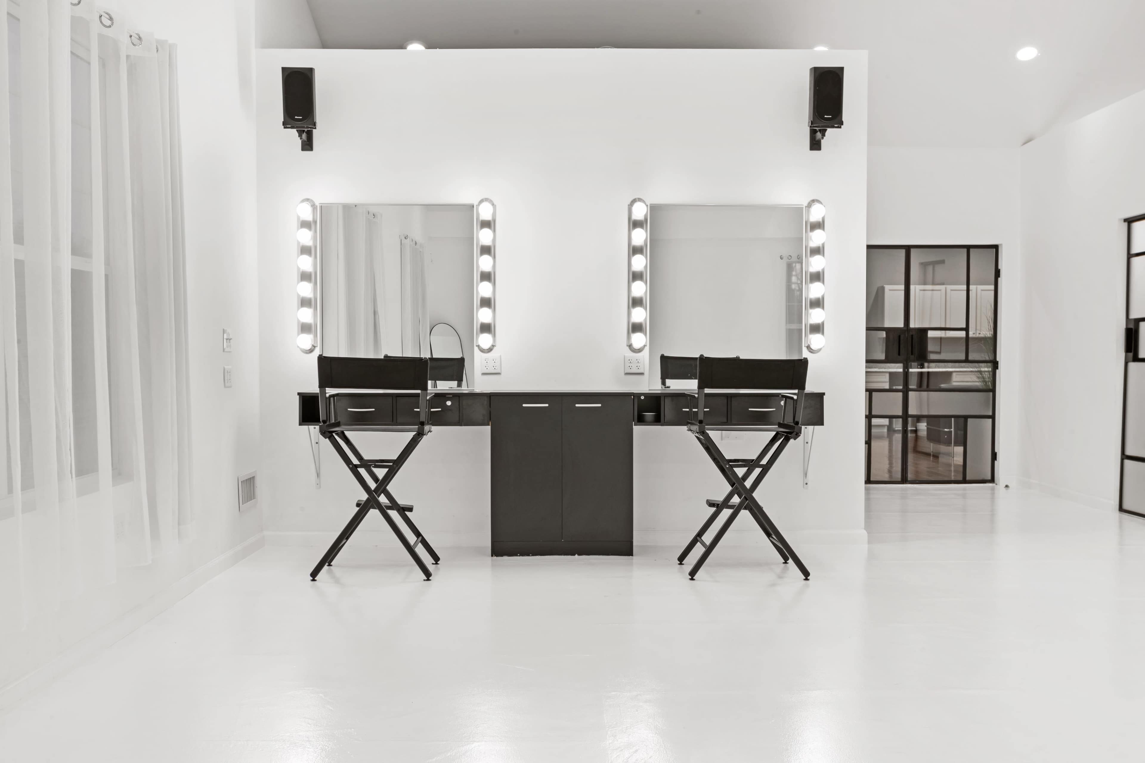 The image shows a modern makeup studio with two black director's chairs positioned in front of illuminated mirrors on a white wall.