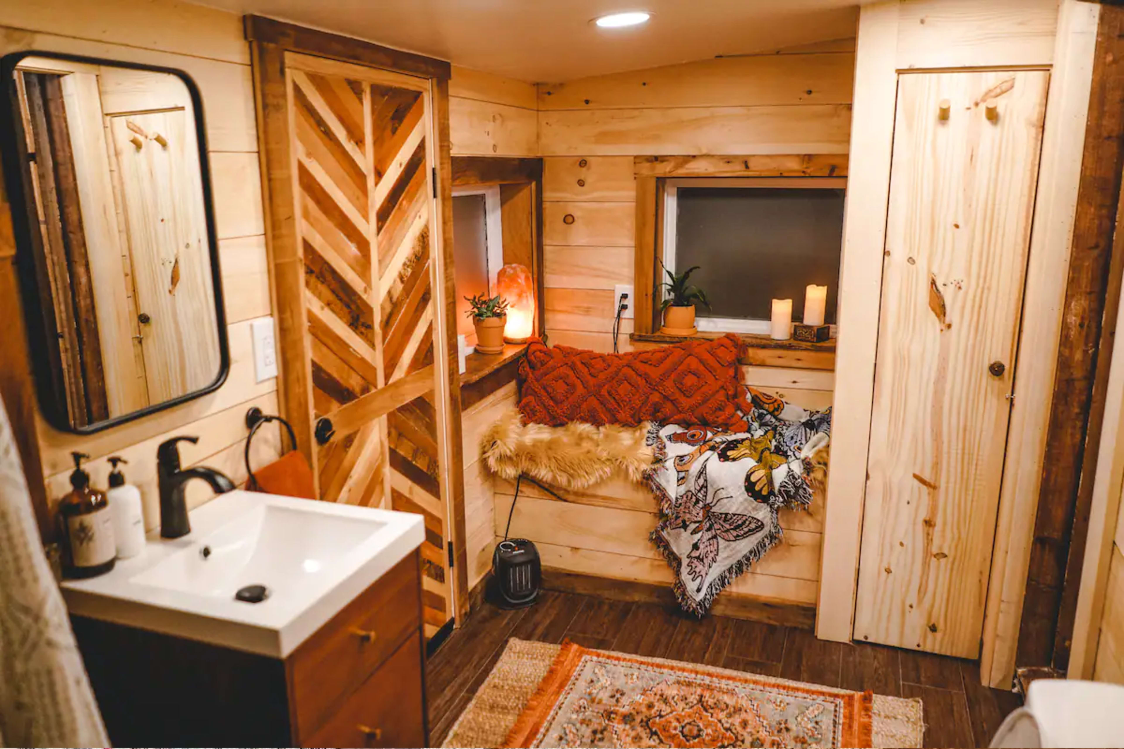 A small, cozy bathroom featuring wooden walls, a mirror, a sink with a dark faucet, a corner seating area with decorative pillows, and soft lighting from candles and a lamp.