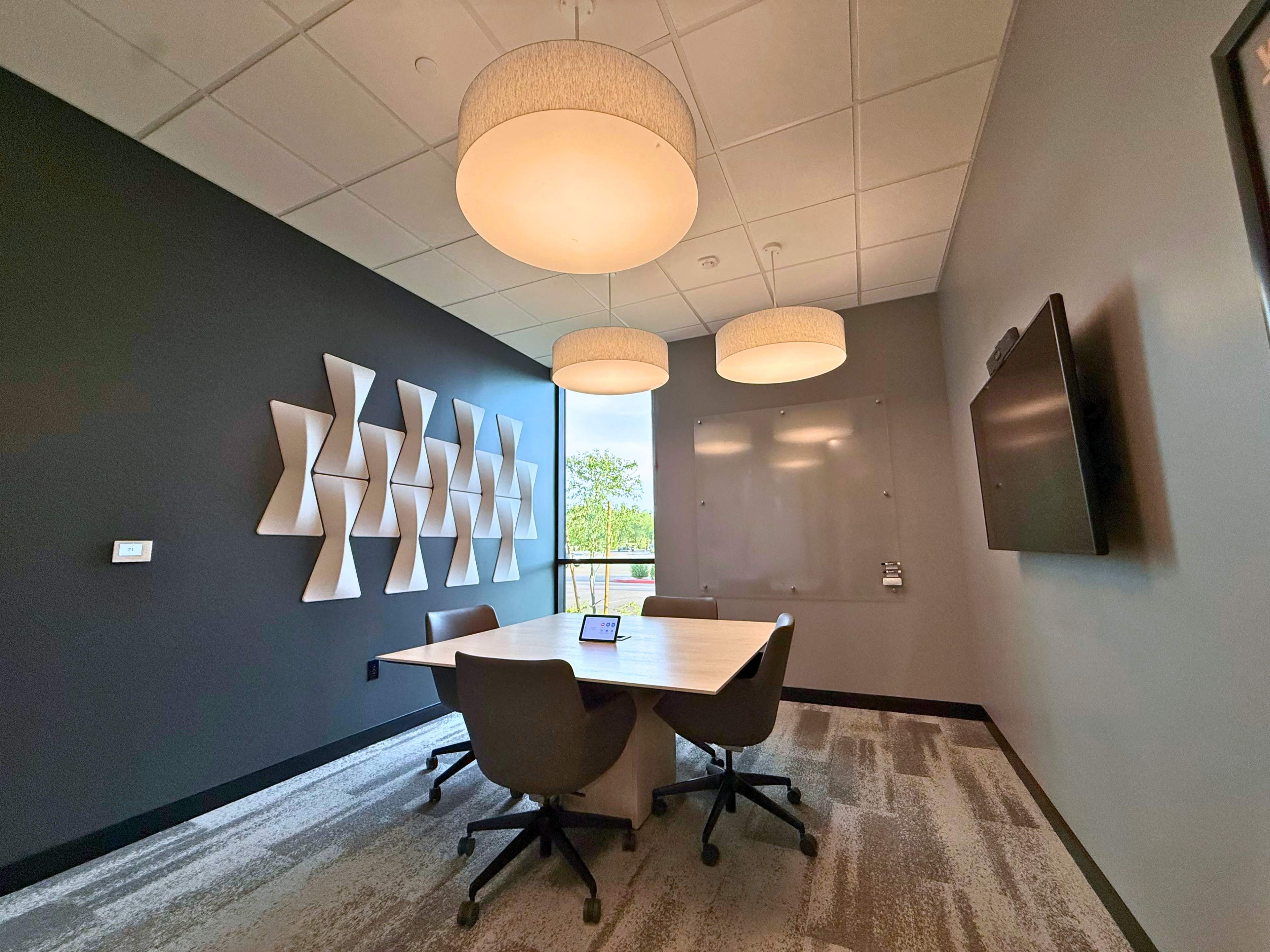A conference room features a large table surrounded by chairs, with modern wall art, two pendant lights, and a mounted screen.