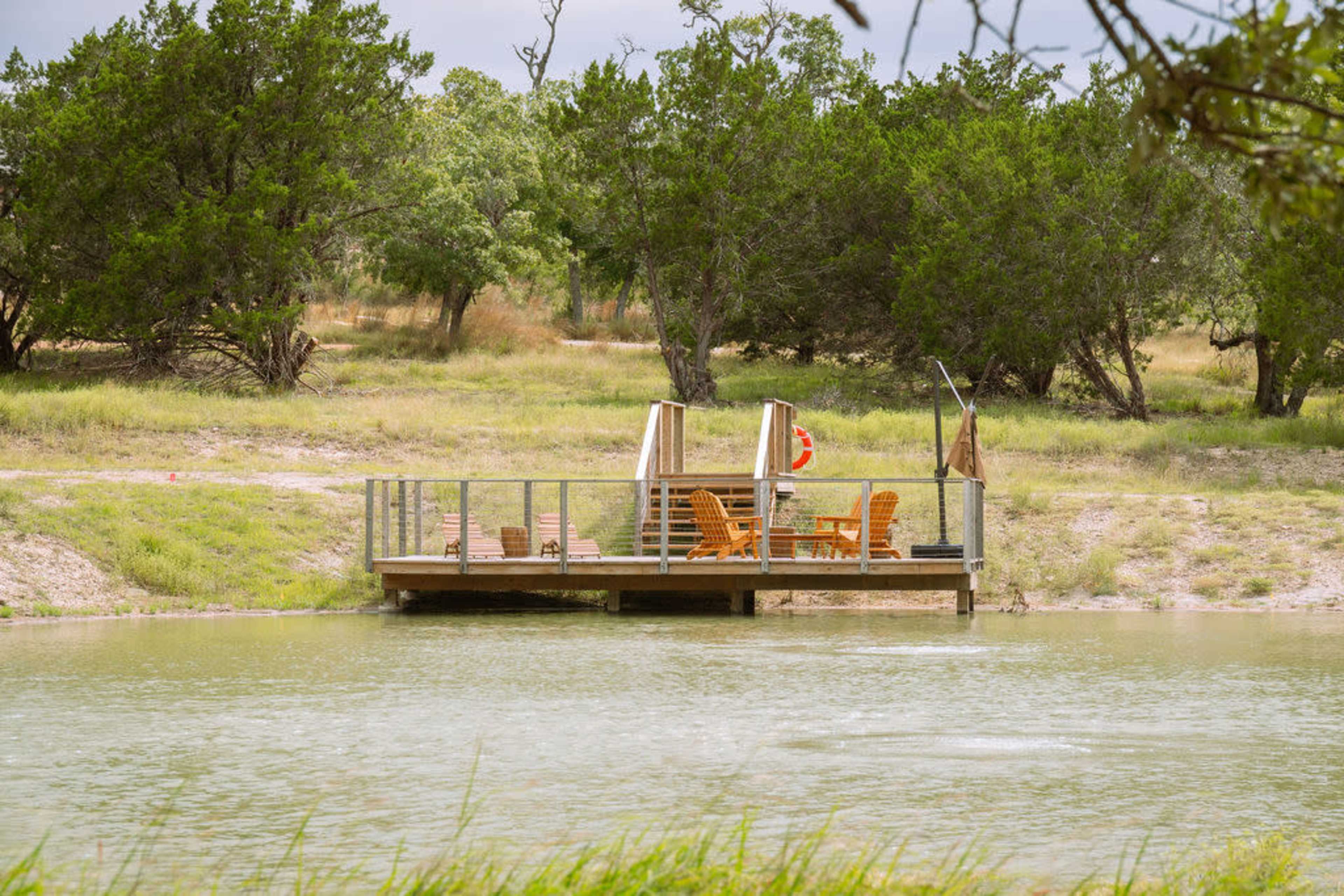 A wooden dock with chairs is situated by a calm pond, surrounded by grass and trees.