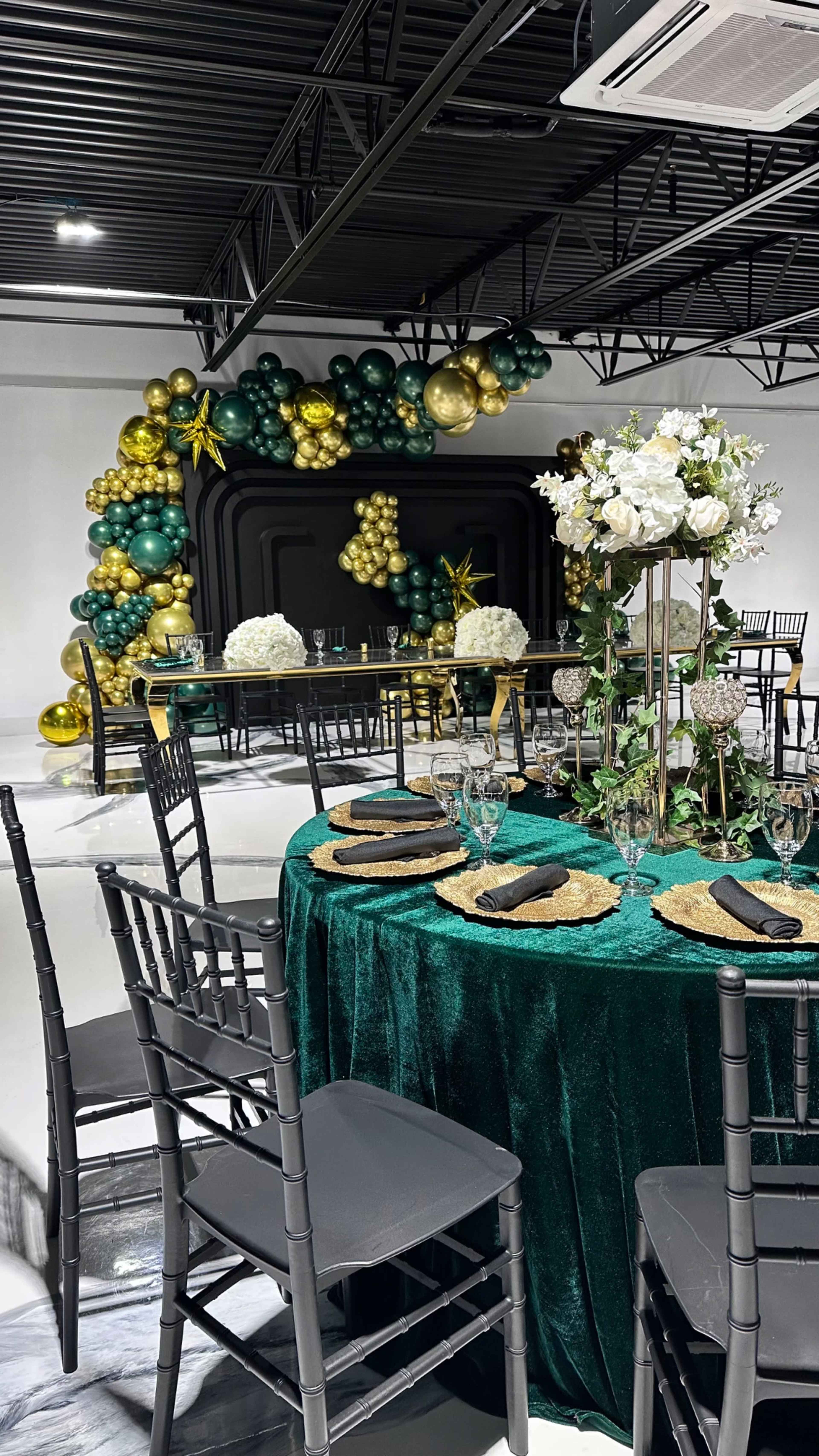 A dining area is set up with a green tablecloth, elegant tableware, and a decorated backdrop featuring green and gold balloons.