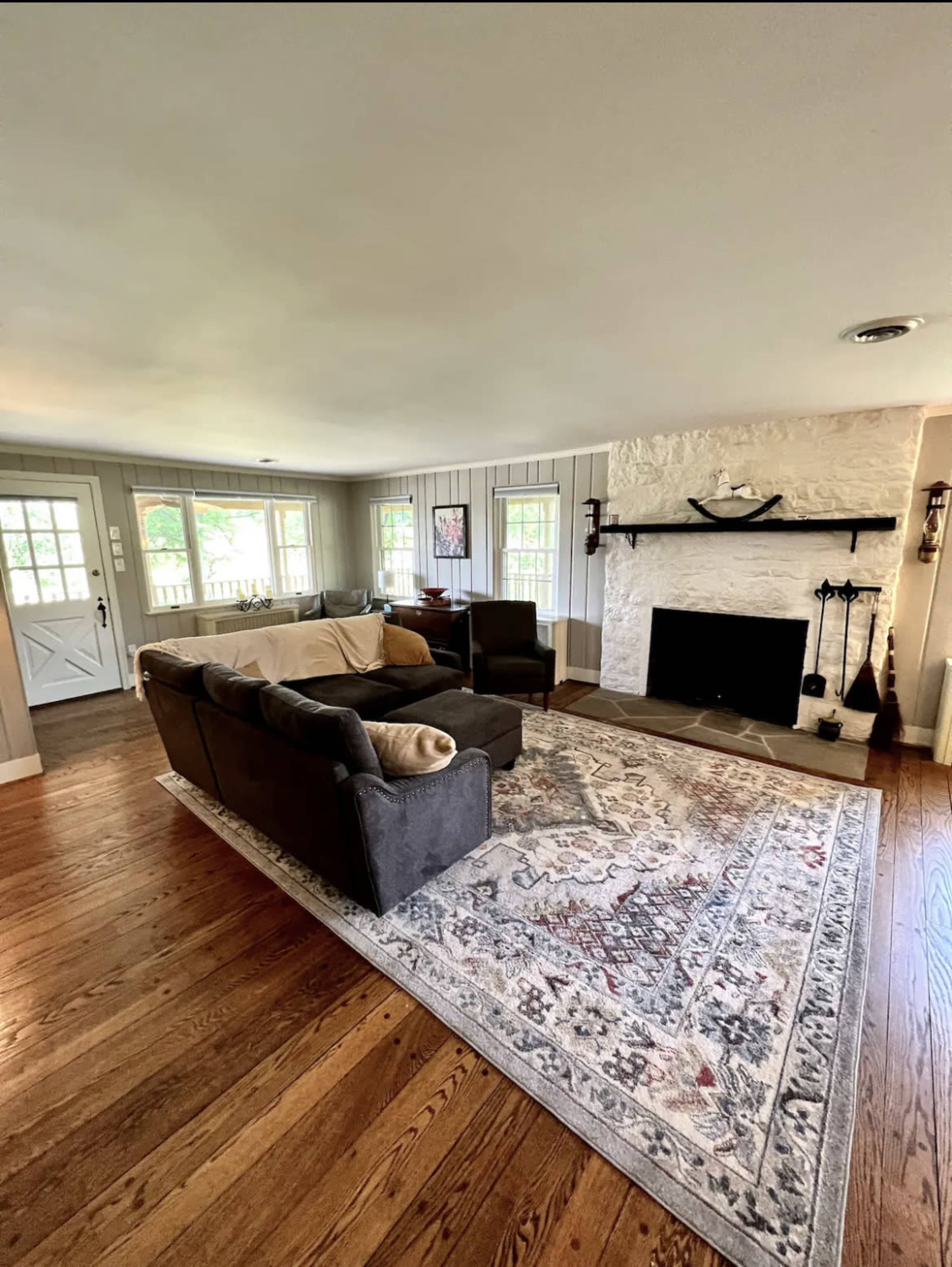 A living room with a light-colored sofa, two armchairs, a large area rug, and a stone fireplace against a white wall.