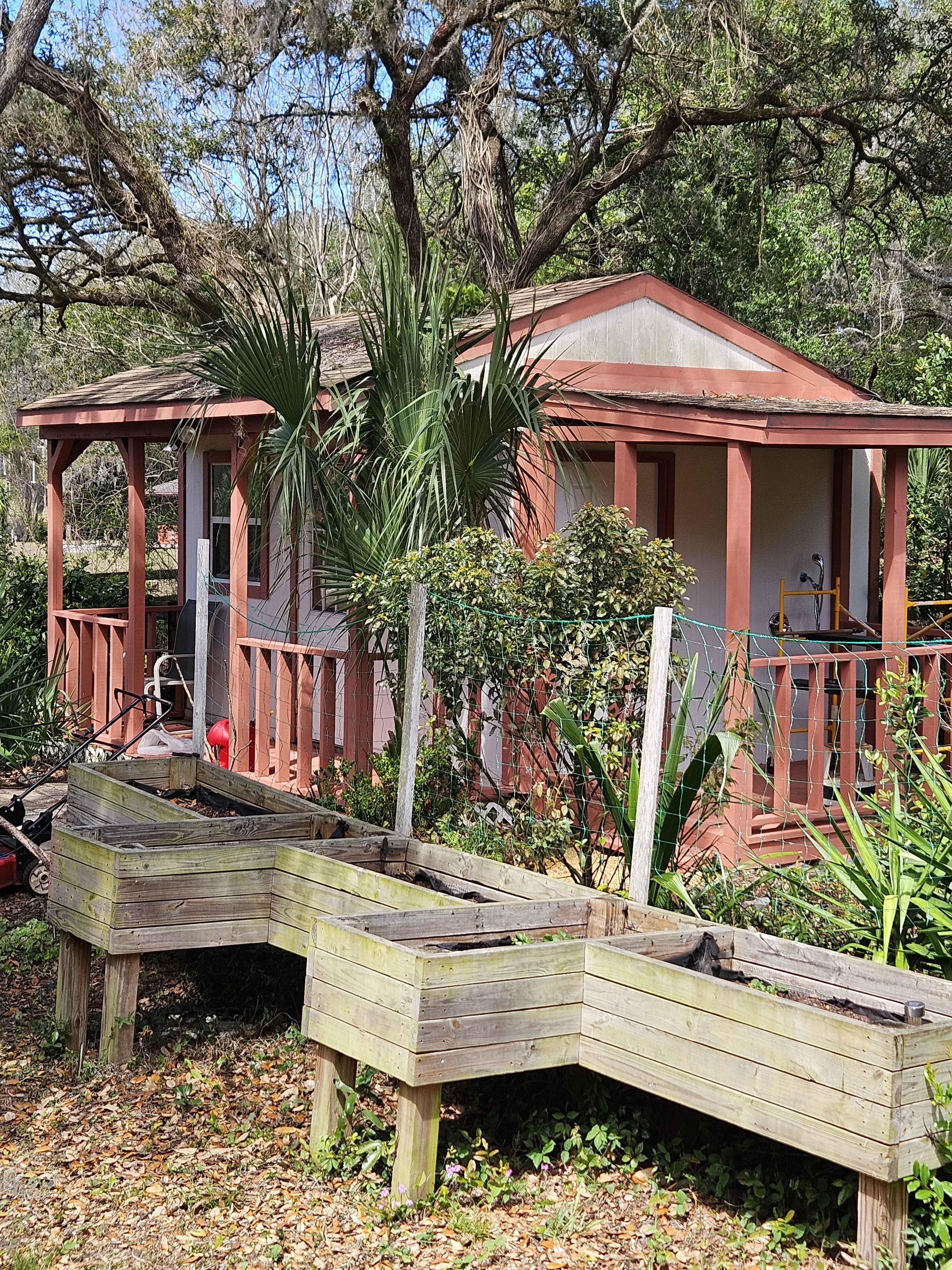 The image shows a small wooden structure with a porch, surrounded by plants and a raised garden bed in front.