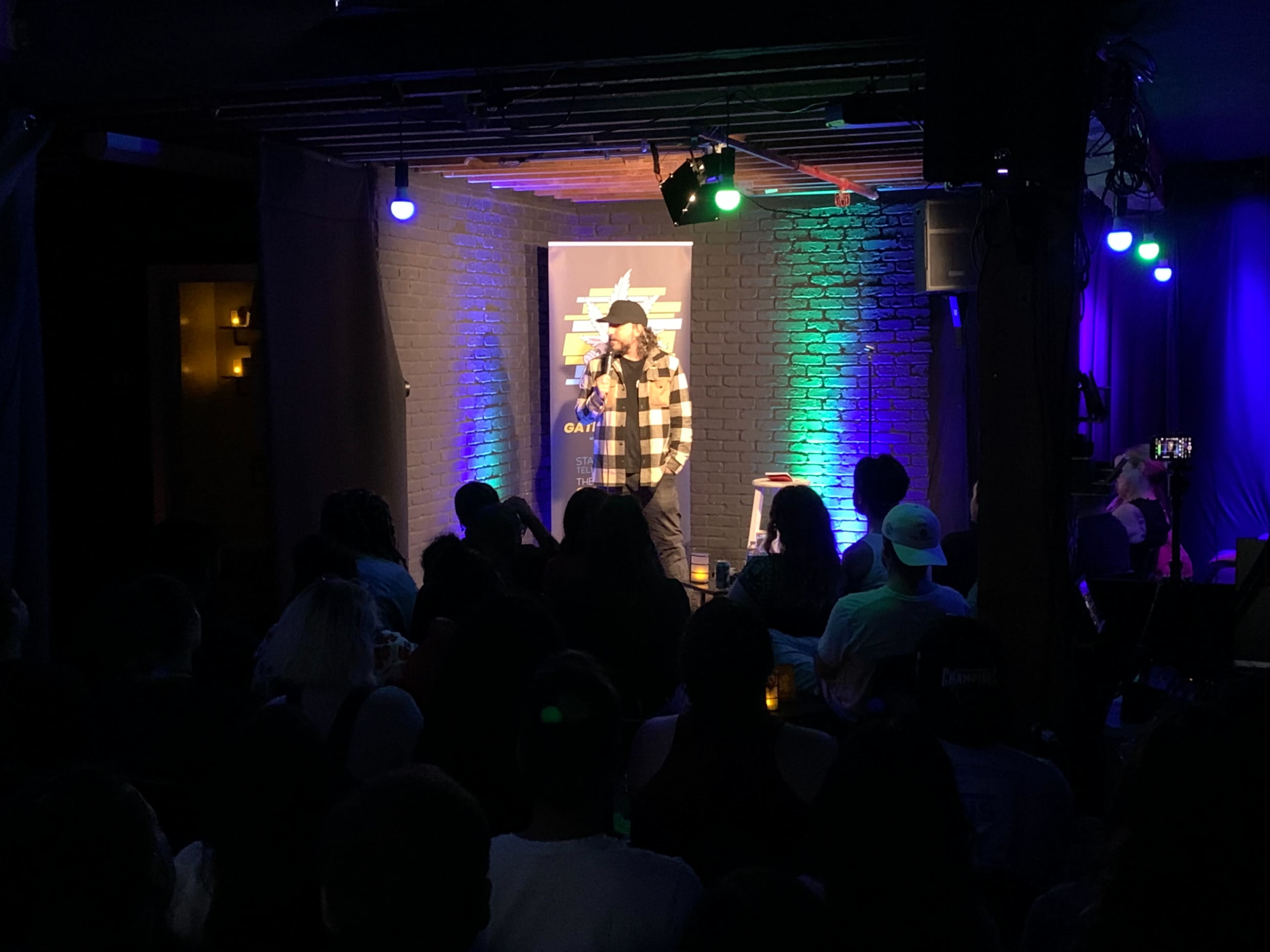 A comedian performs on stage in a dimly lit venue with a crowd seated in front of him.