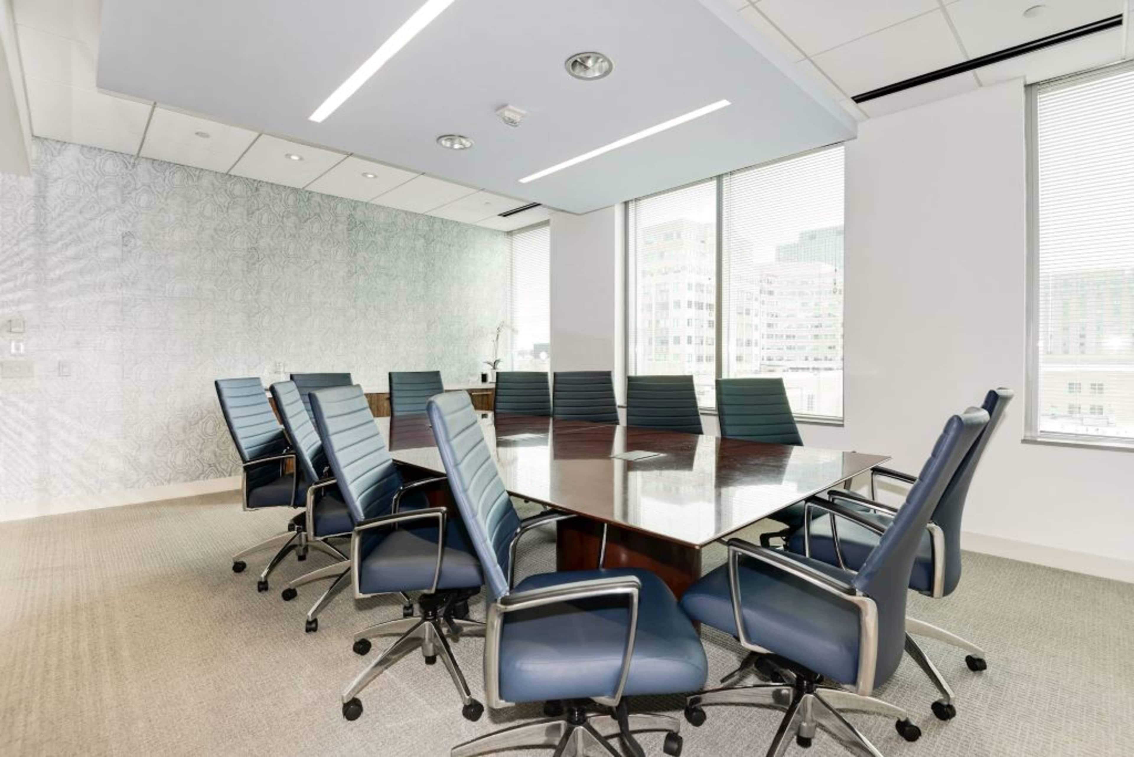 A conference room features a large wooden table surrounded by ergonomic office chairs and large windows allowing natural light.