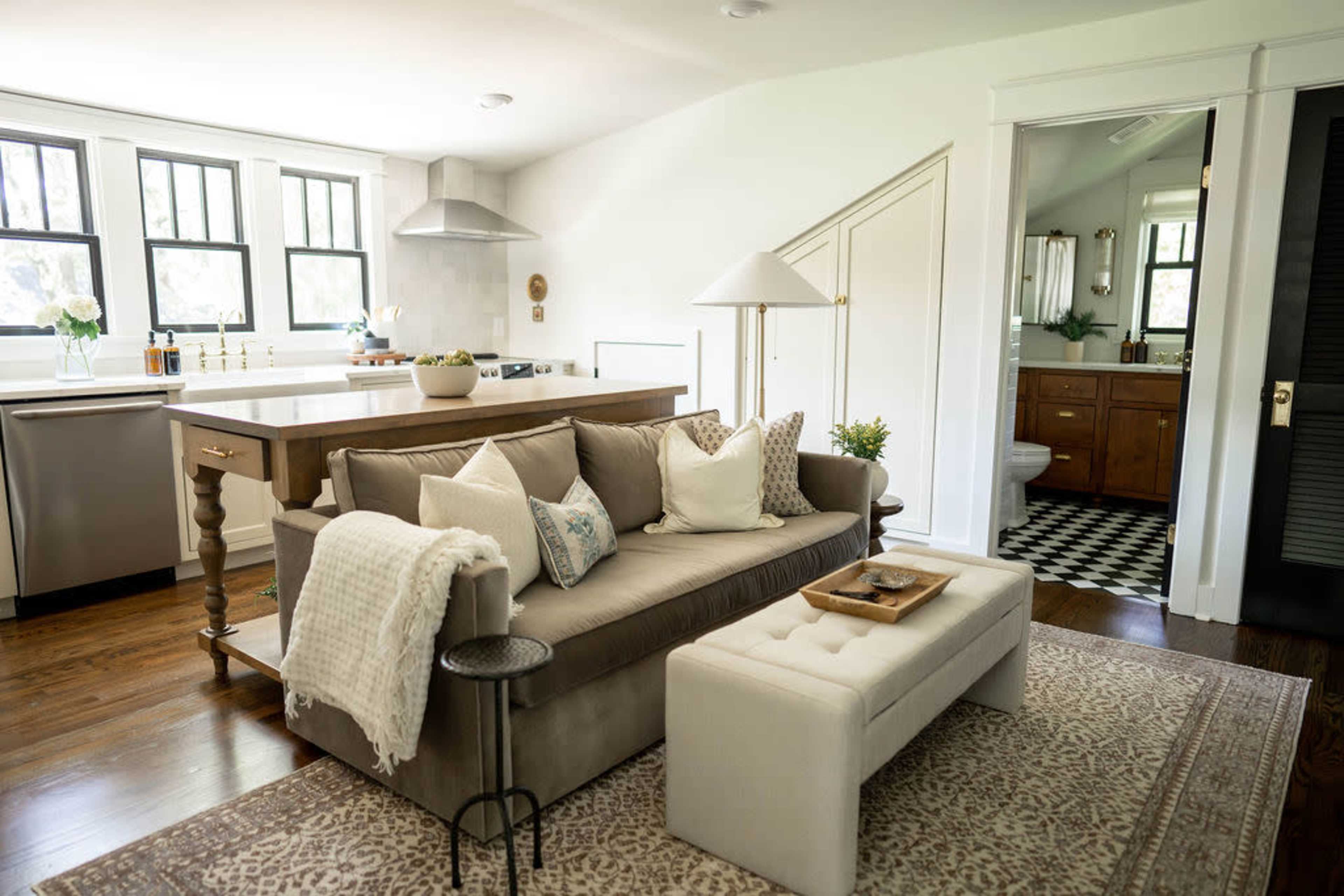 A light-filled living room featuring a beige sofa with decorative pillows, a white ottoman, and a kitchen area in the background with a wooden island.