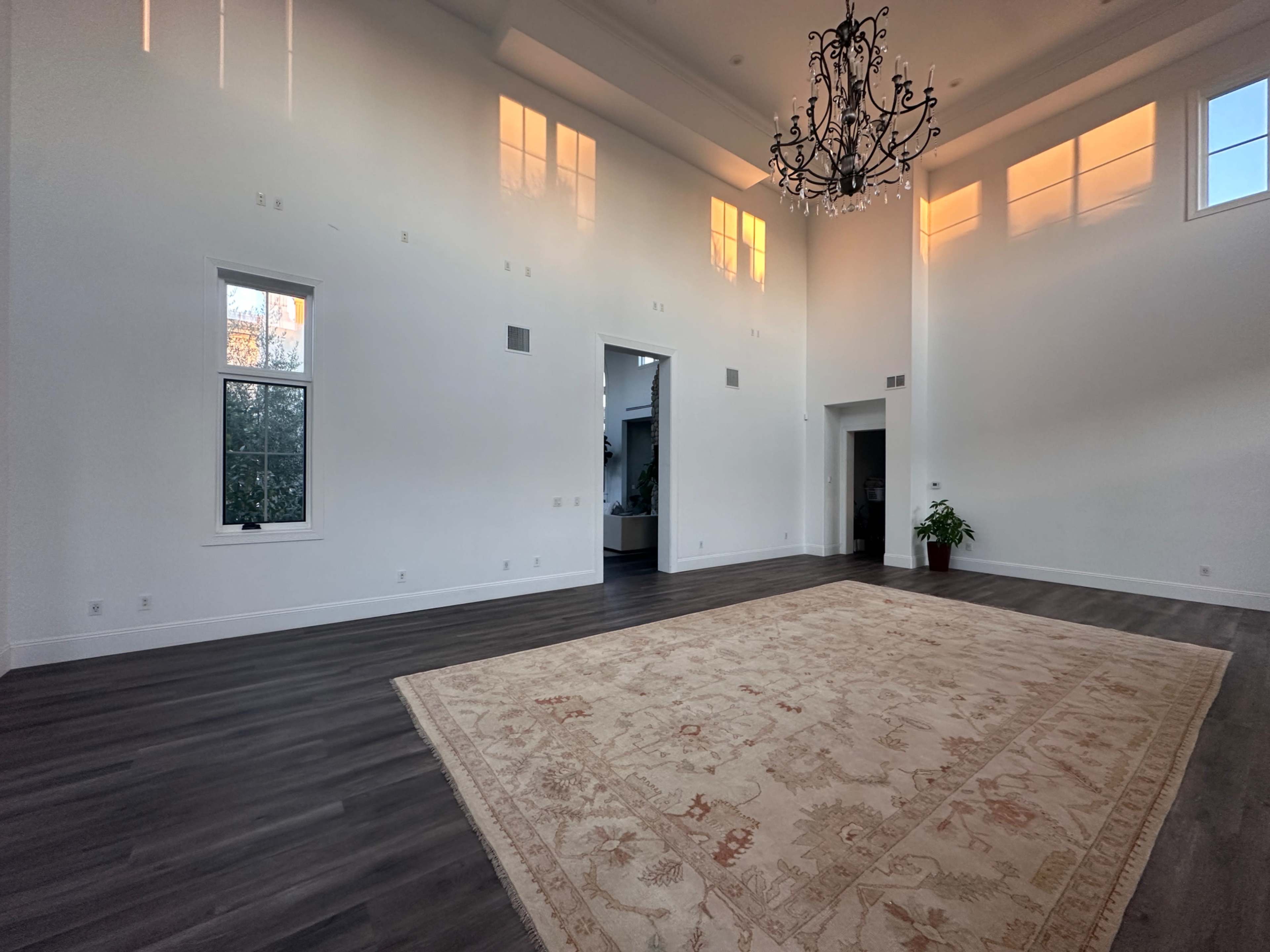The image shows a spacious, well-lit room with a large patterned rug in the center, tall windows displaying a sunset, and a chandelier hanging from the ceiling.