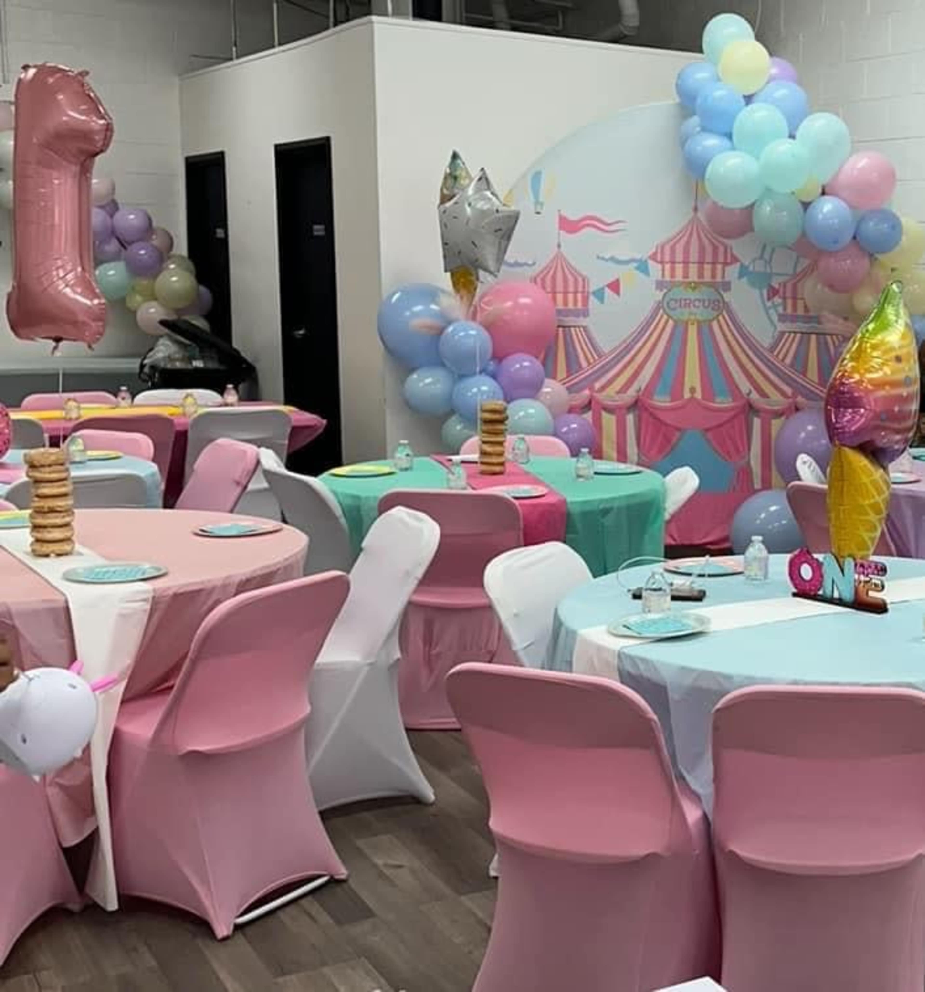 The scene shows a decorated party space with tables covered in pink and white linens, balloons in various colors, and a circus-themed backdrop featuring a colorful tent.