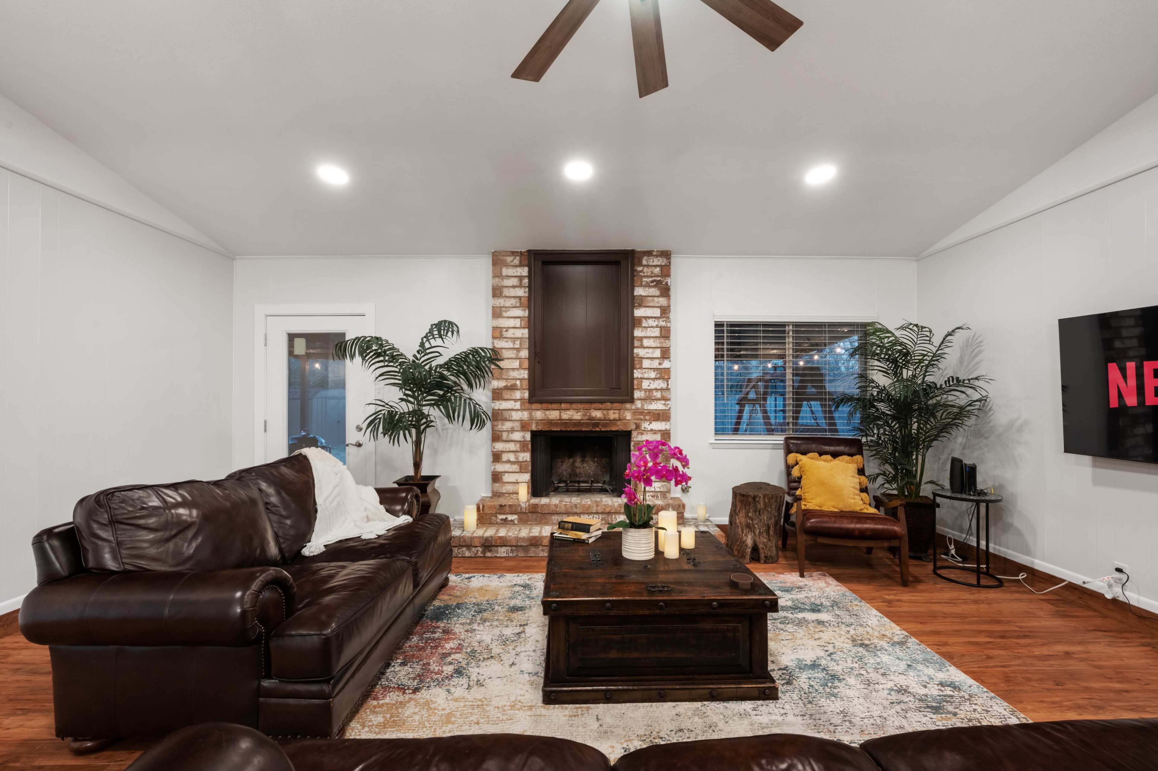 The living room features a brown leather sofa, a brick fireplace, and a coffee table set on a patterned rug, with plants and decorative items enhancing the space.