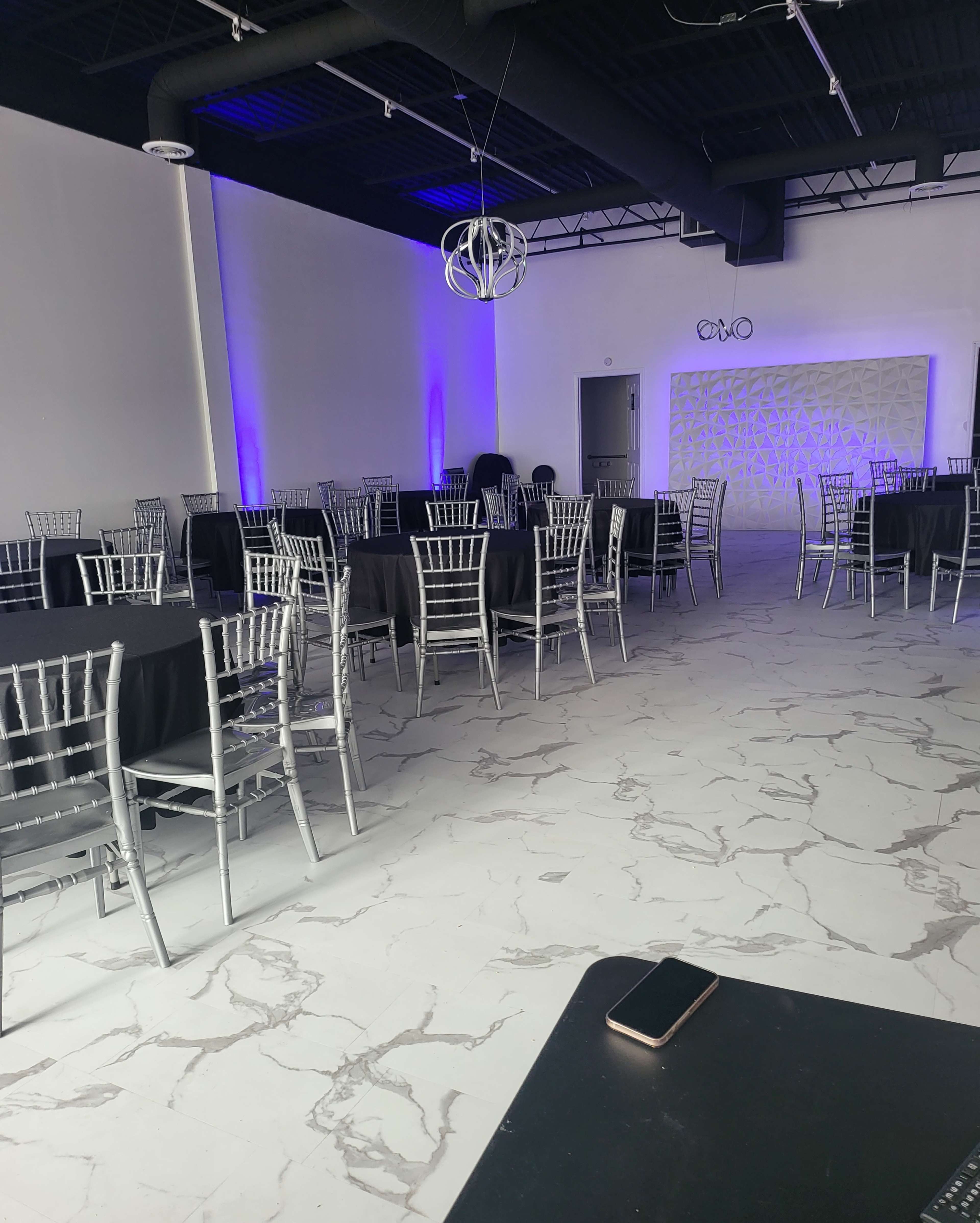 A spacious event venue filled with silver chiavari chairs and black tables arranged throughout the room, featuring purple lighting and a textured wall at one end.