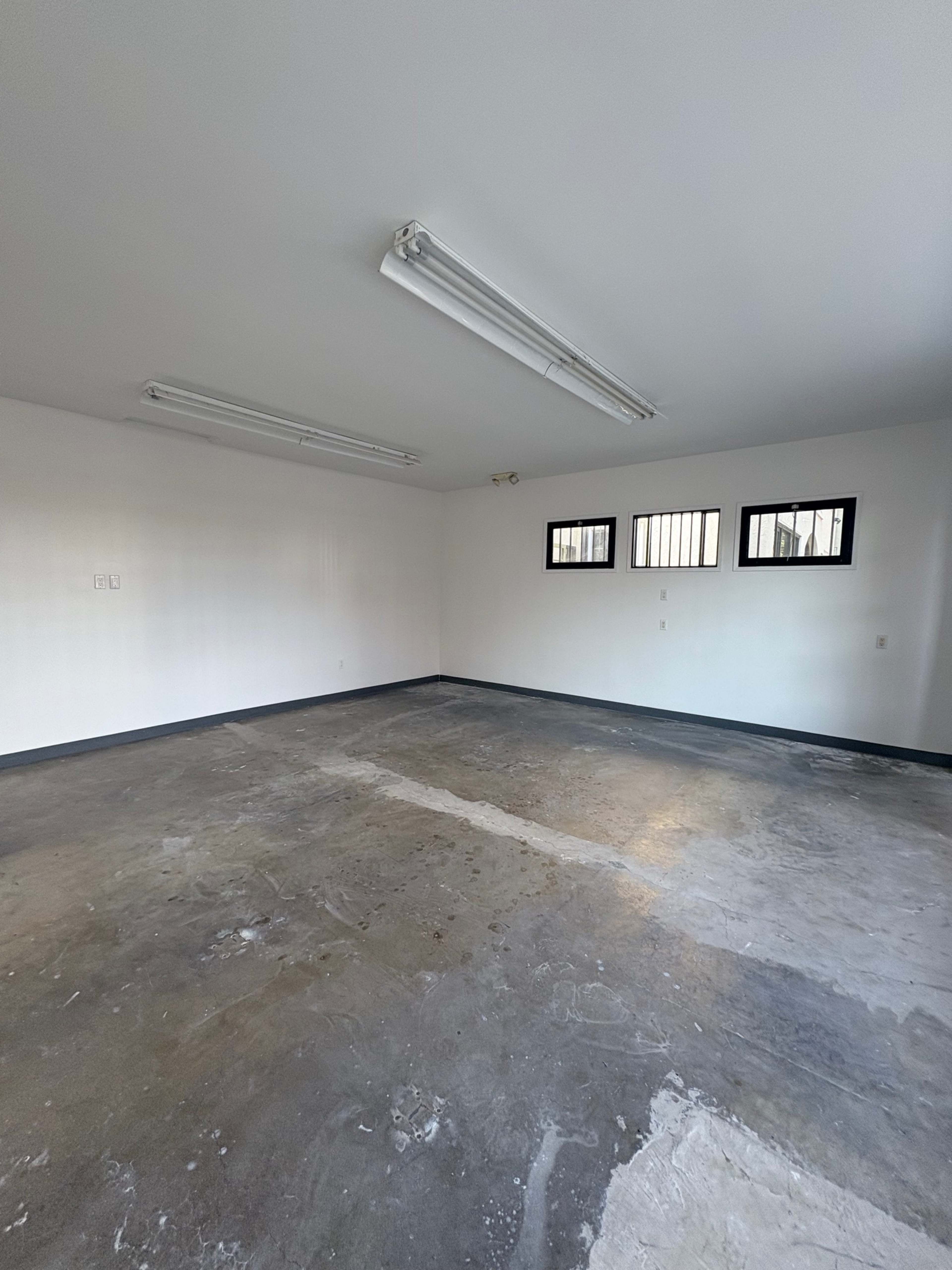 The image shows an empty room with a concrete floor, white walls, and three small windows.