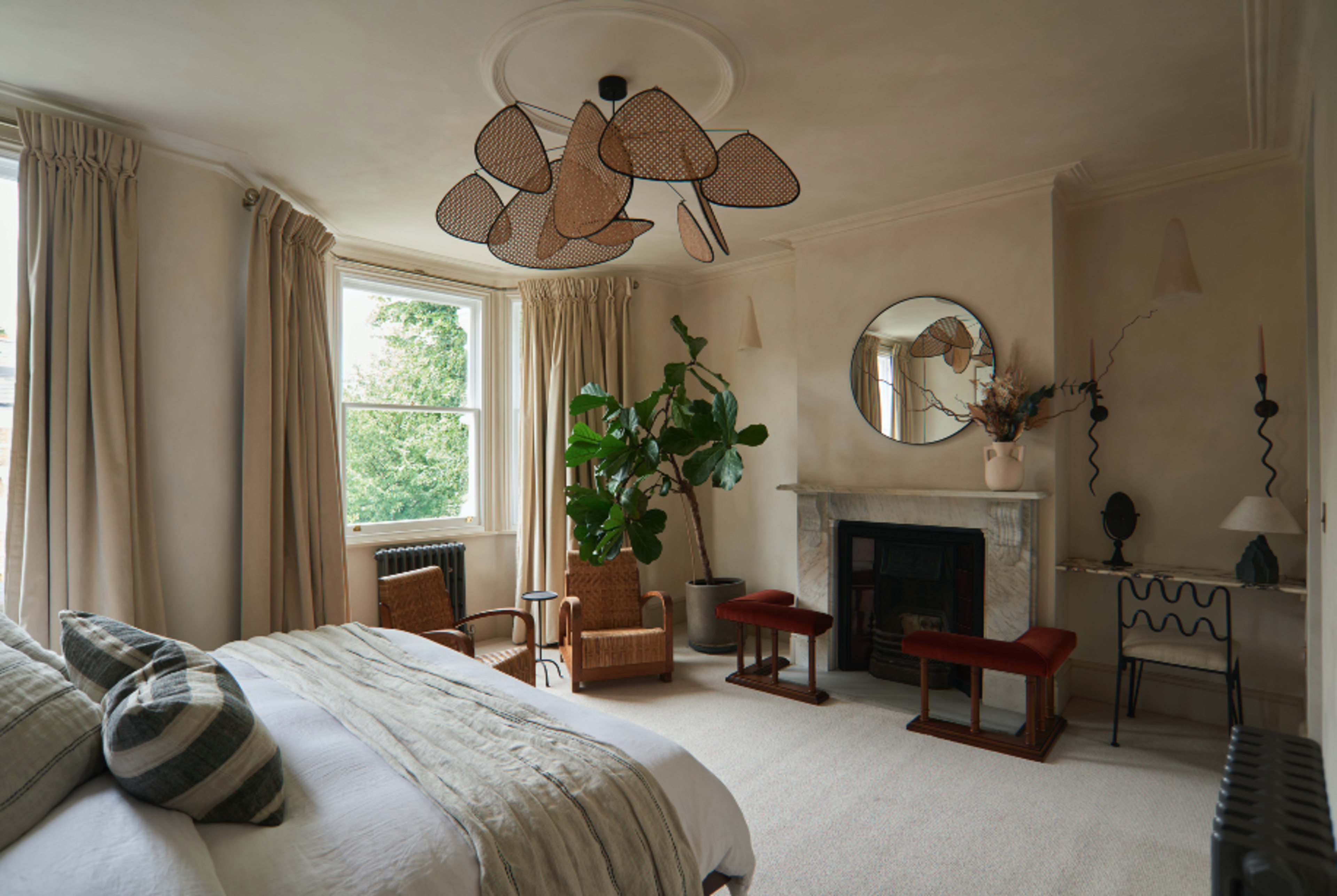 The room features a bed with neatly arranged linens, a large window with curtains, a plant in the corner, and a fireplace framed by decorative elements.