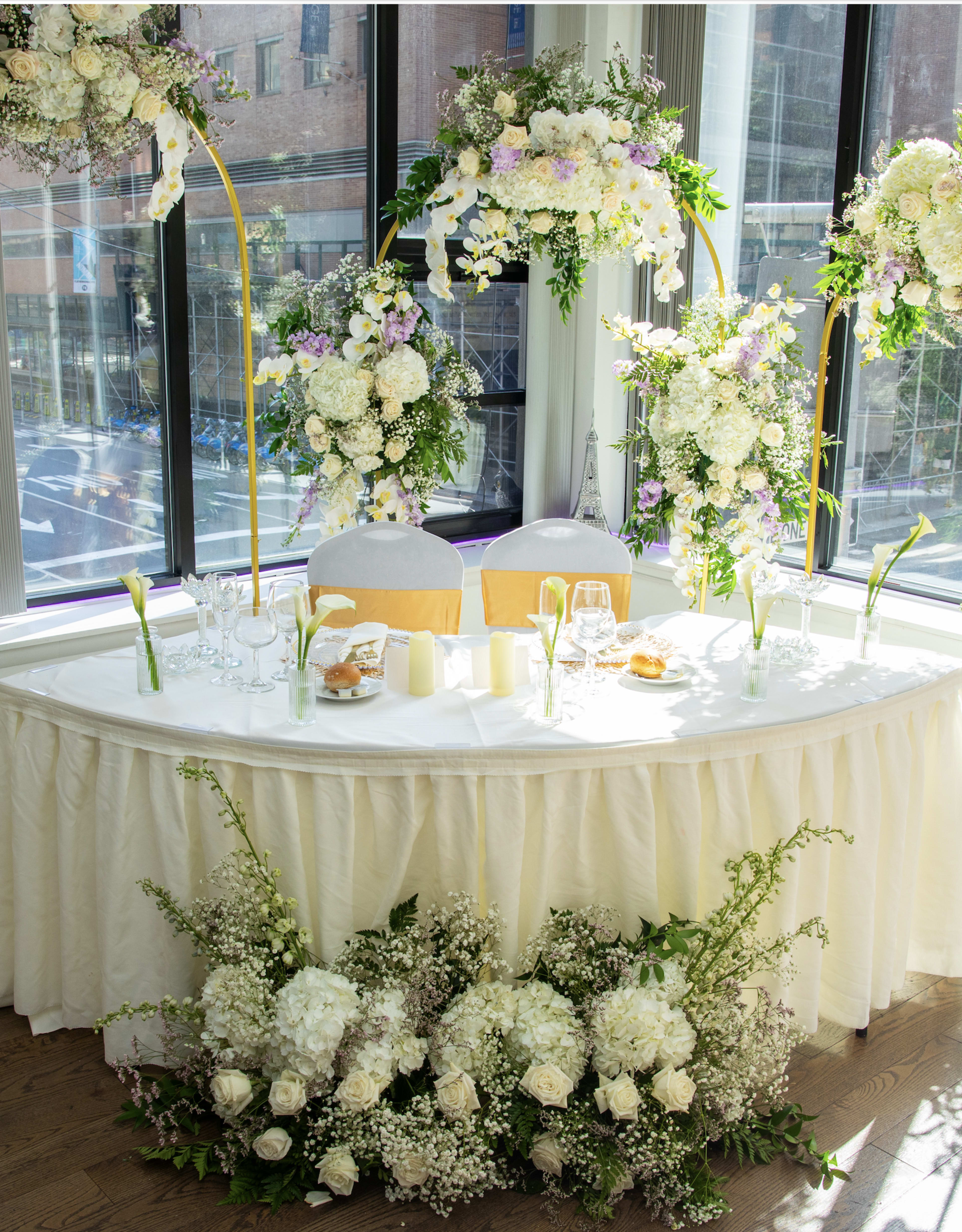A beautifully decorated wedding table is set against large windows, adorned with floral arrangements and candles, featuring white and pastel flowers.