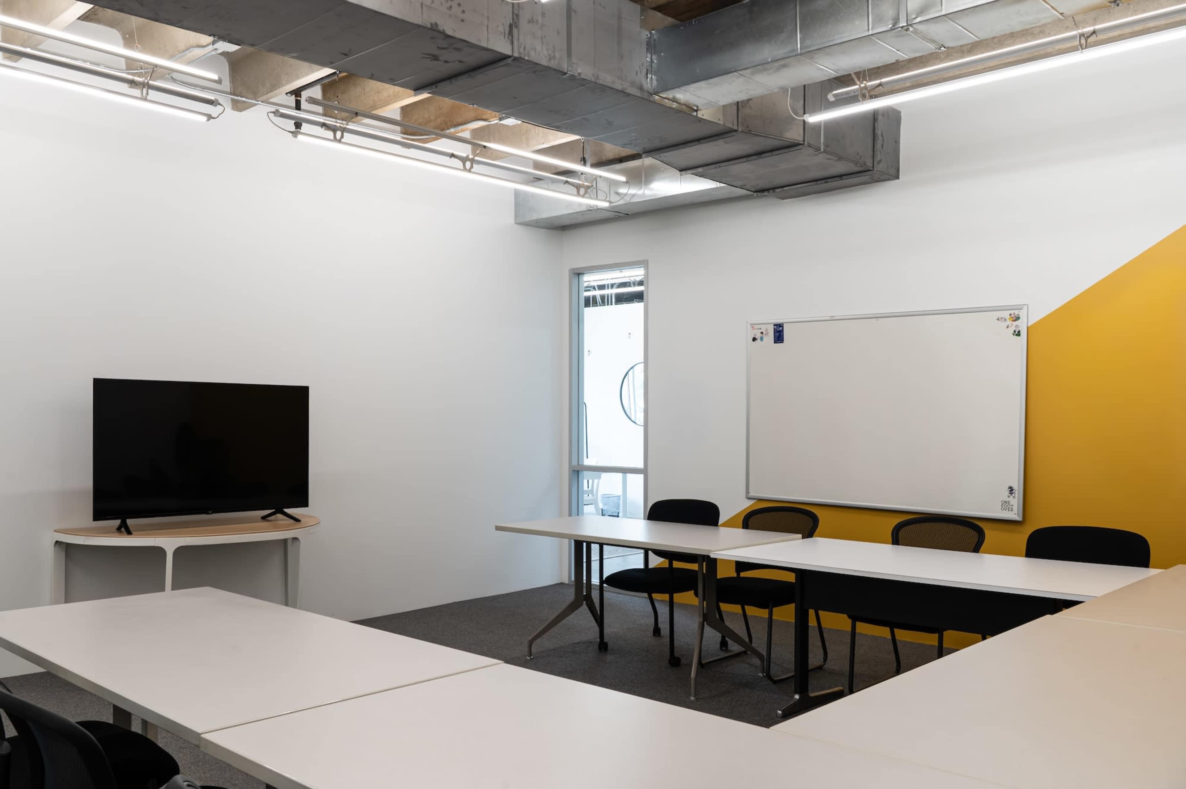 The image shows a modern conference room with a large television screen, a whiteboard, and several tables arranged in a U-shape, complemented by a yellow accent wall.