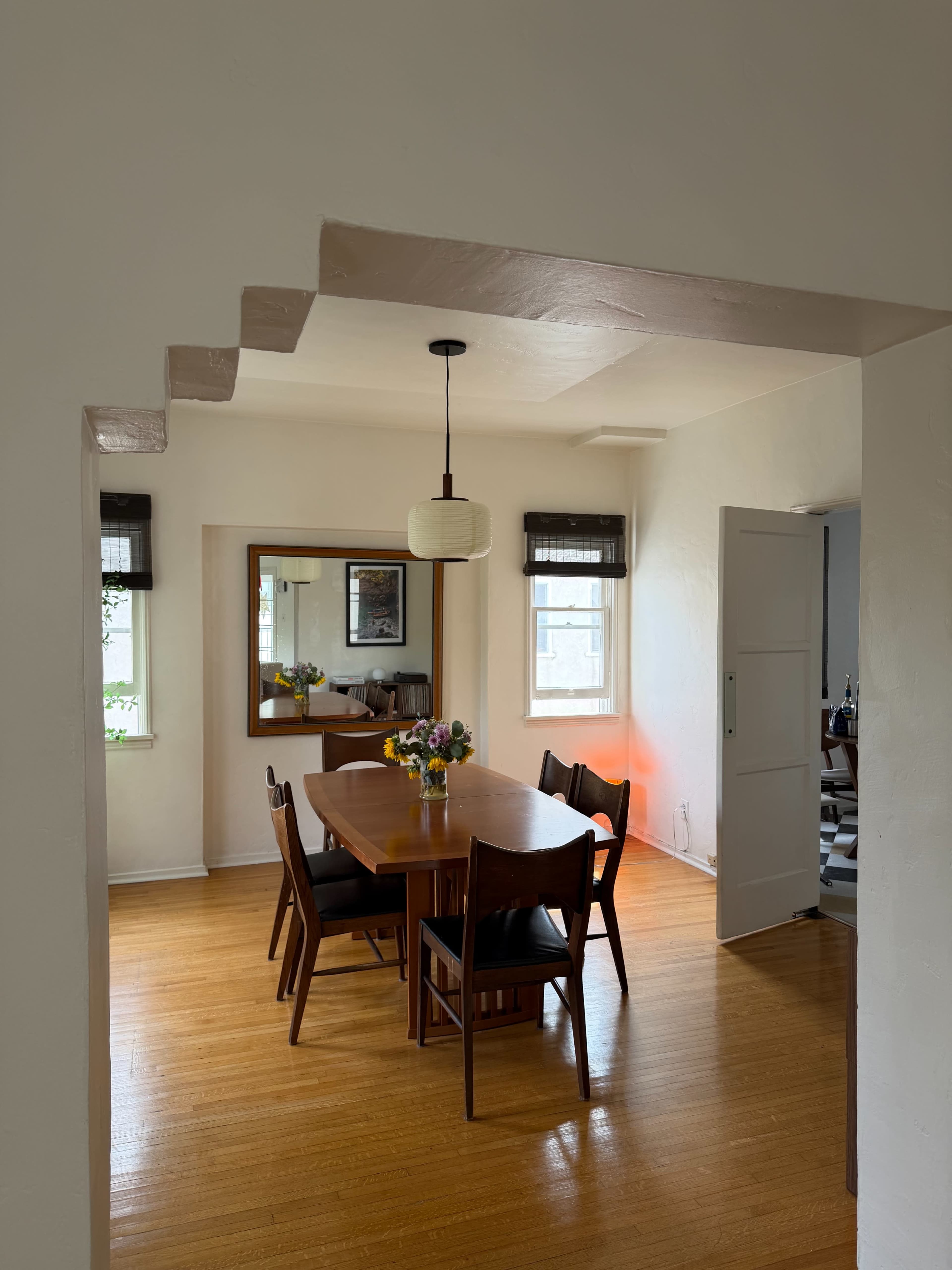 A dining area features a wooden table surrounded by chairs, with a pendant light hanging above and a mirror on the wall.