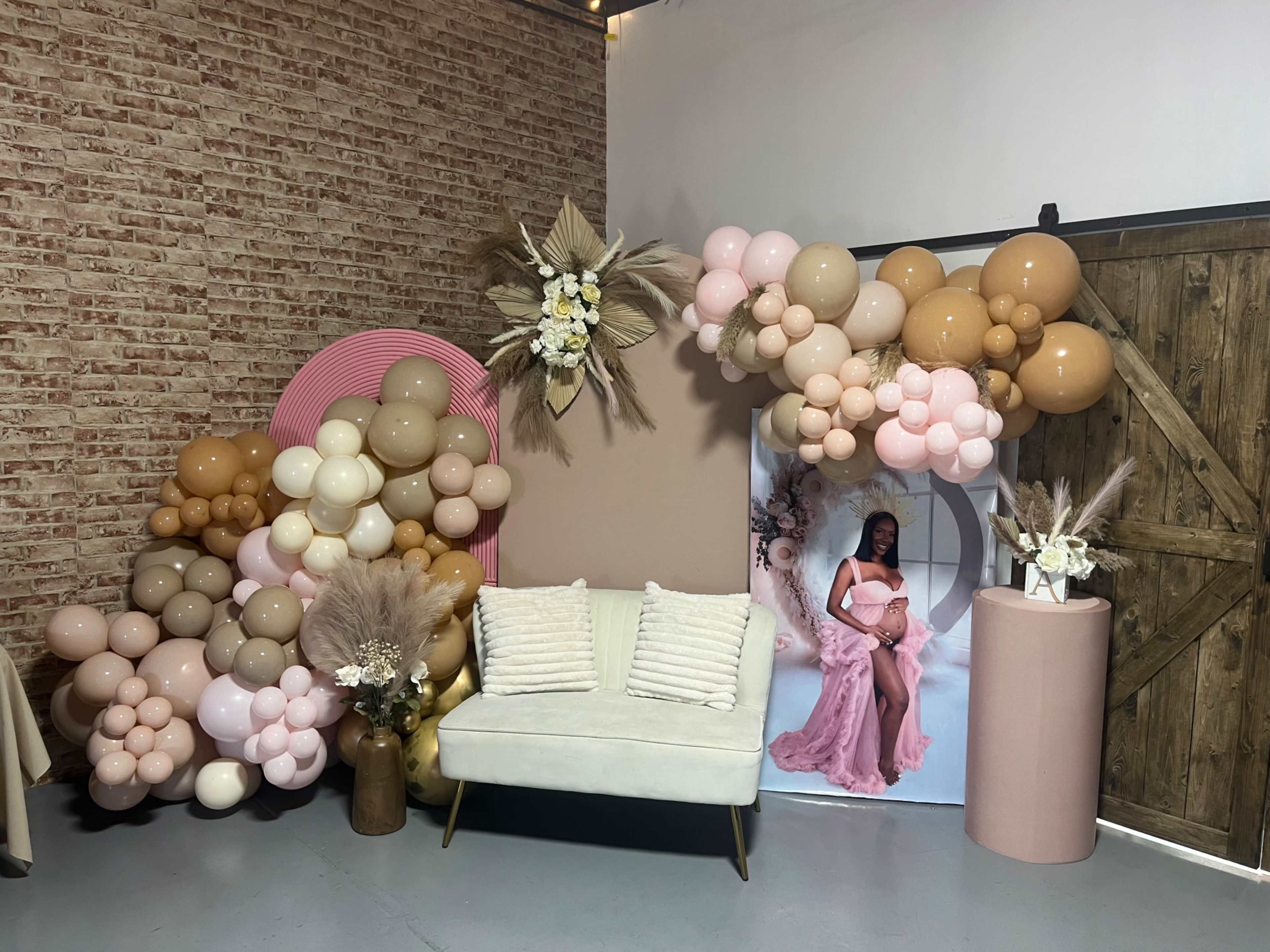 A decorated corner with a backdrop of balloons, a floral arrangement, a round pink display, and a white couch in front of a large photo of a woman in a pink gown.