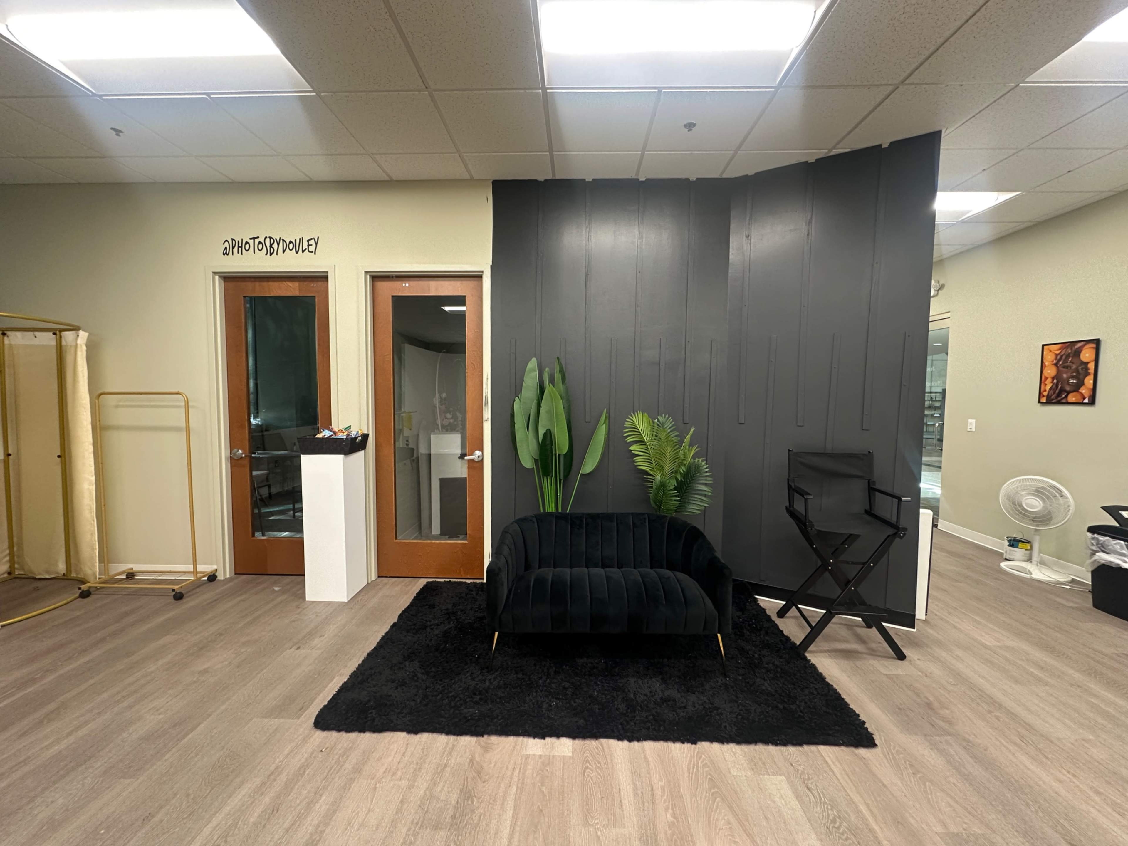 The image shows a modern office space featuring a black velvet couch, a plant, and a black rug, with two glass doors leading to another area.