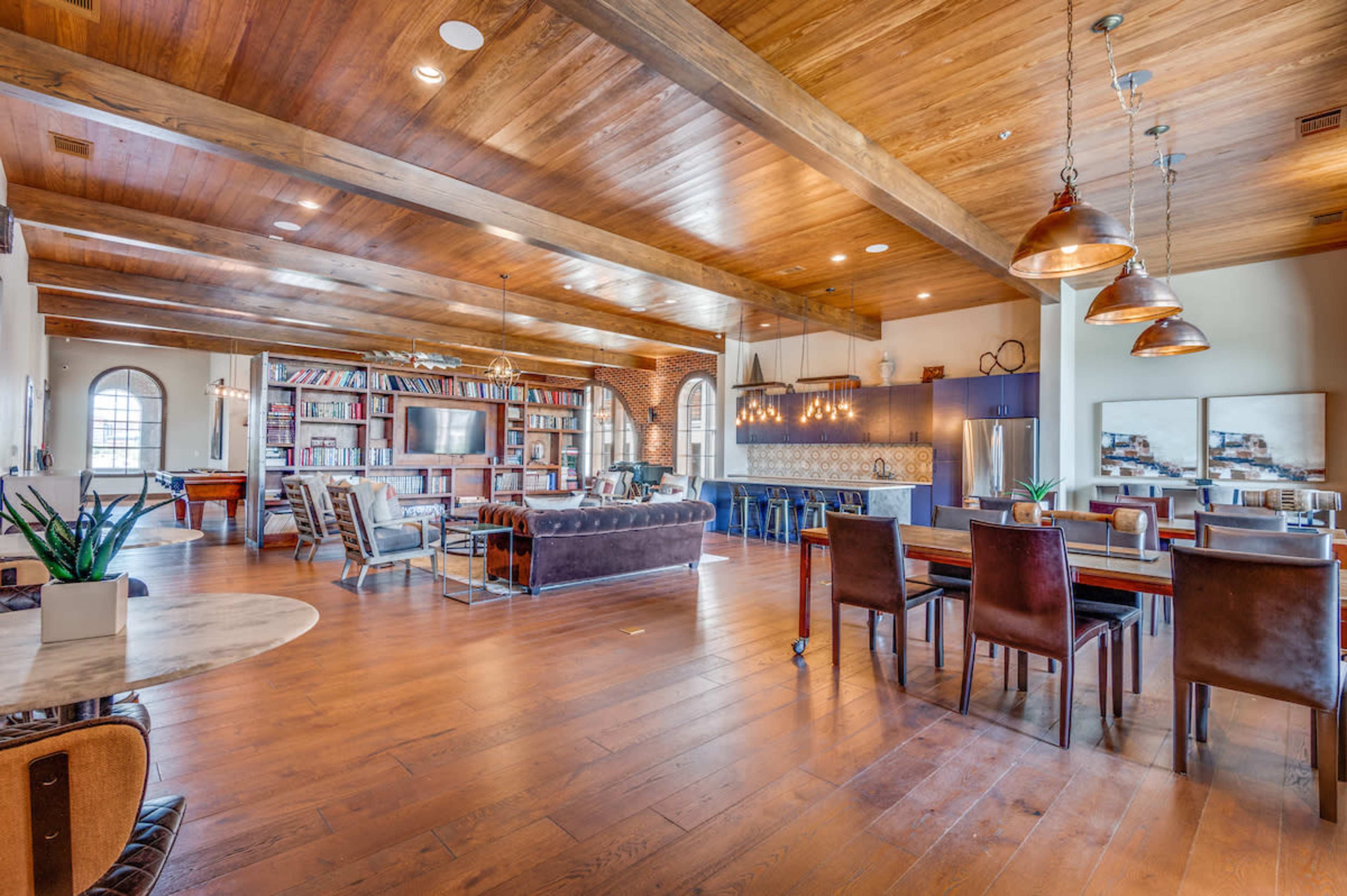 The spacious room features wooden beams and flooring, divided into a living area with a couch and bookshelf, and a dining area with a table and chairs.
