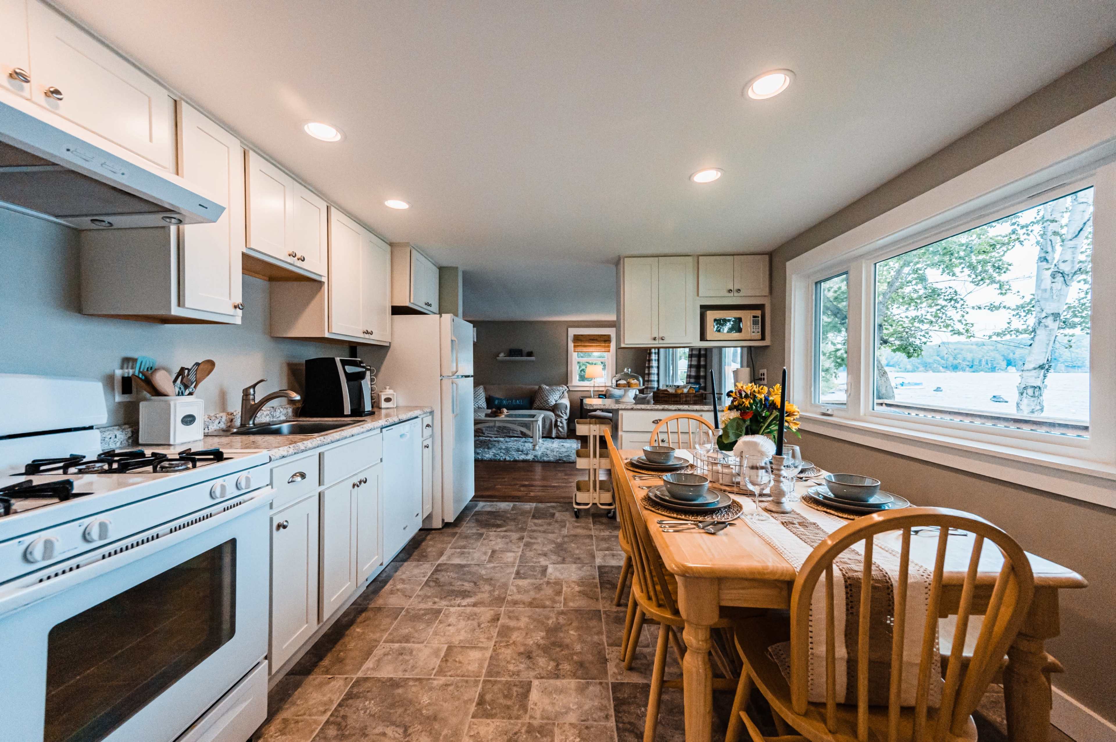 A neatly arranged modern kitchen with white cabinetry, a dining table set for four, and a view of a cozy living area through a large window.