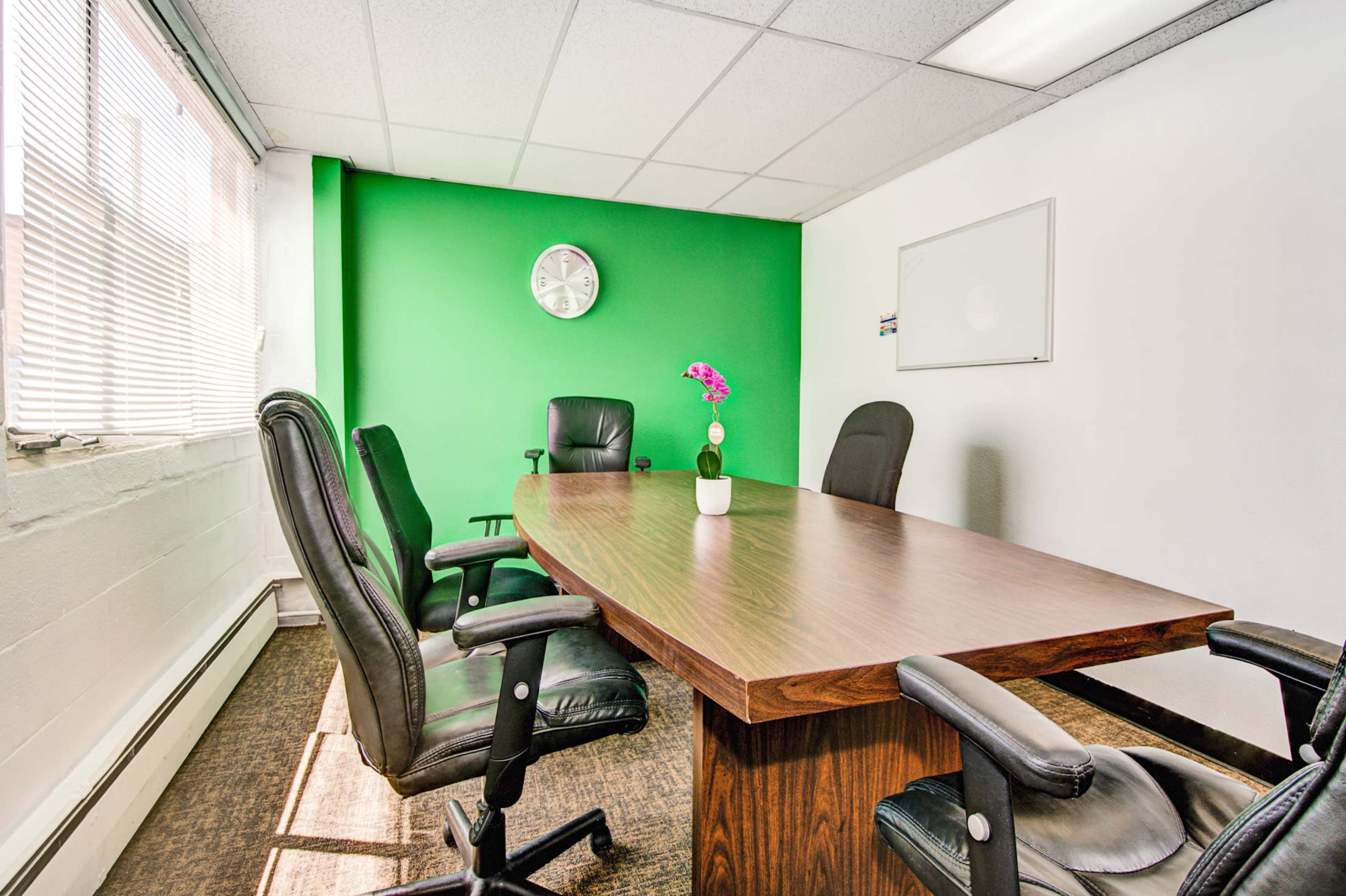 A conference room features a long wooden table surrounded by black office chairs, with a green accent wall and a clock mounted on the wall.