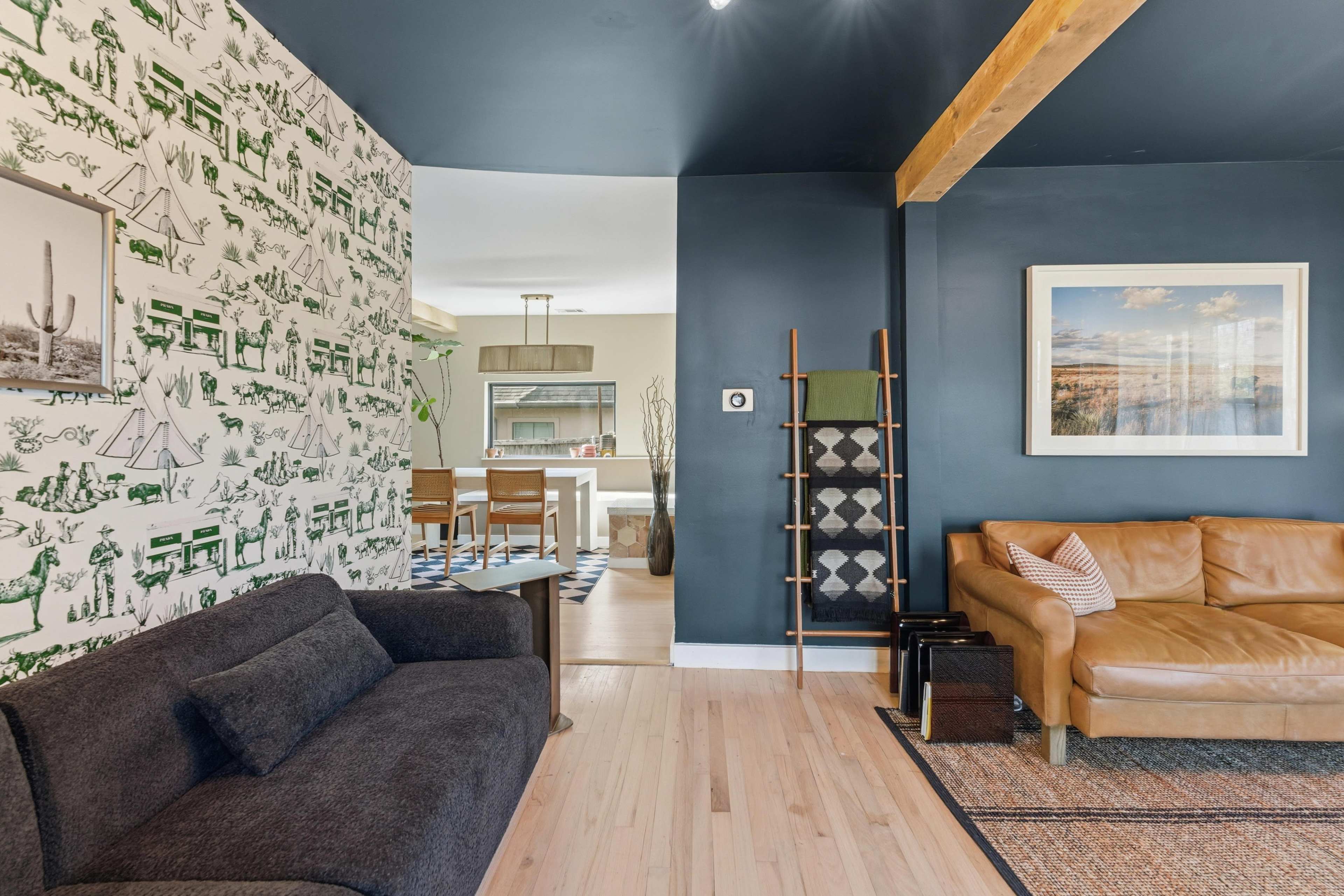 The image shows a living room featuring a dark blue wall, a patterned wallpaper depicting a desert scene, a brown leather sofa, and a dining area visible in the background.