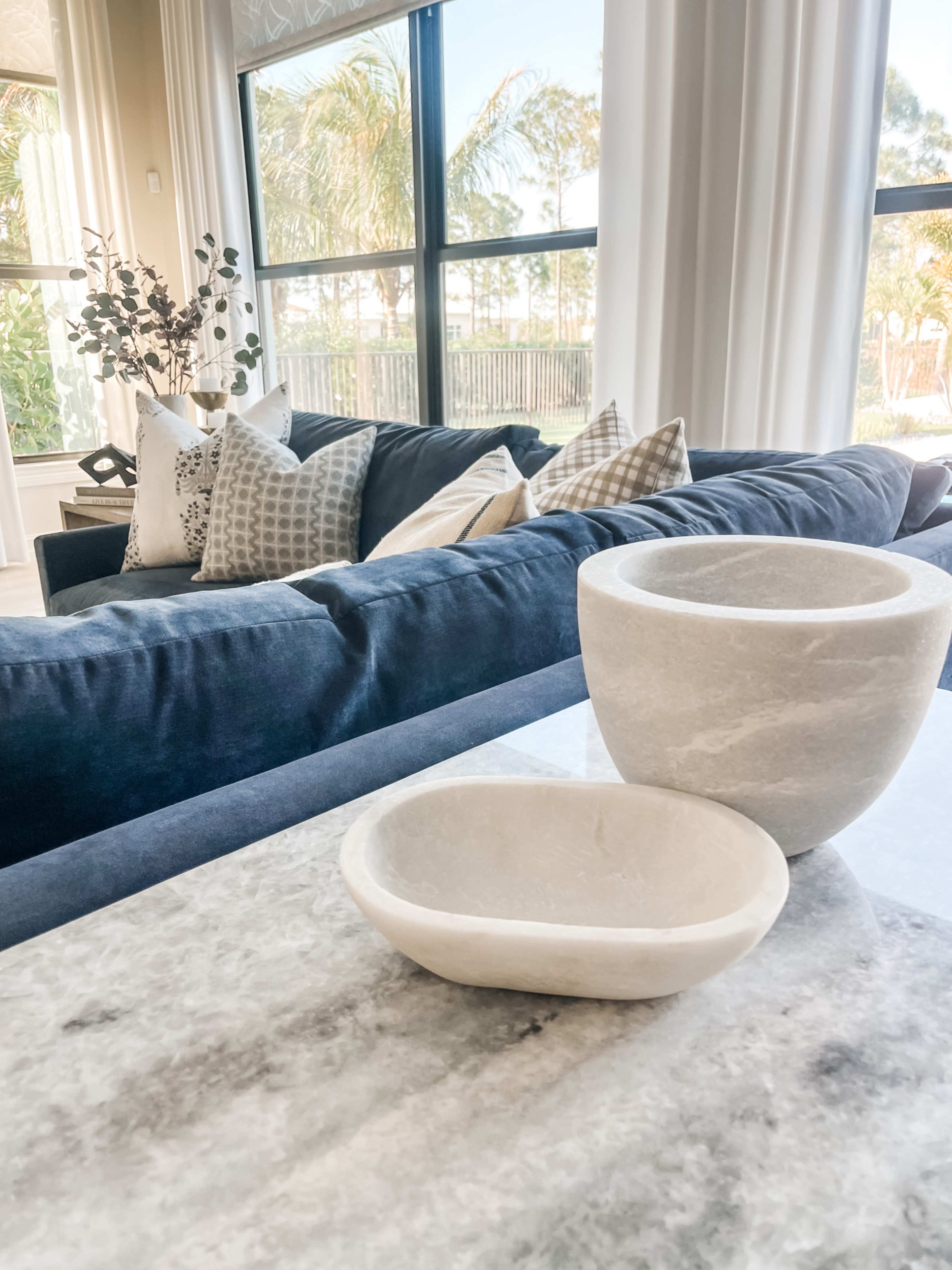 A marble tabletop displays two white stone bowls in front of a dark blue sofa adorned with patterned pillows.