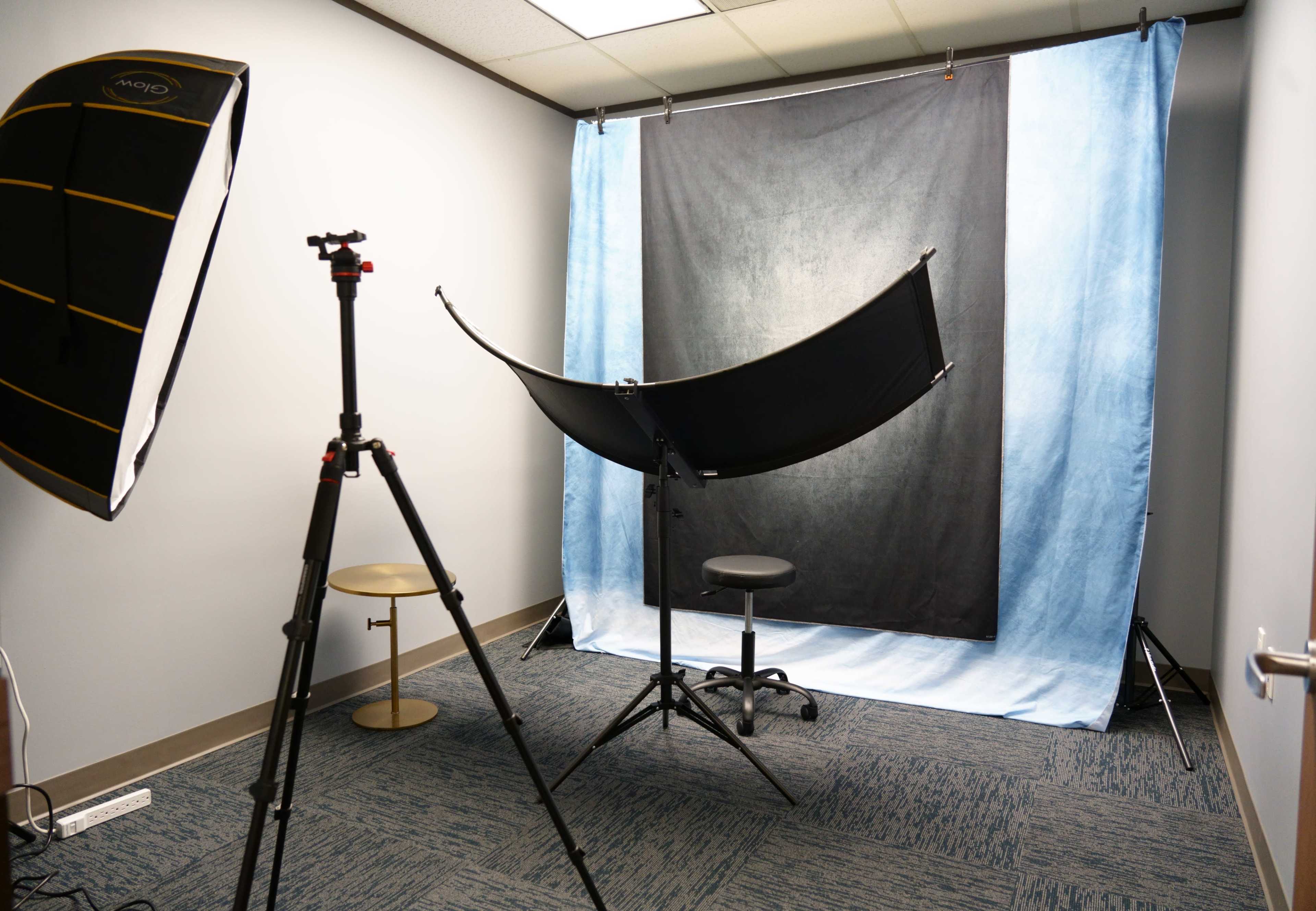 The image shows a photo studio setup with a black and blue backdrop, a tripod, a light source, and a stool.