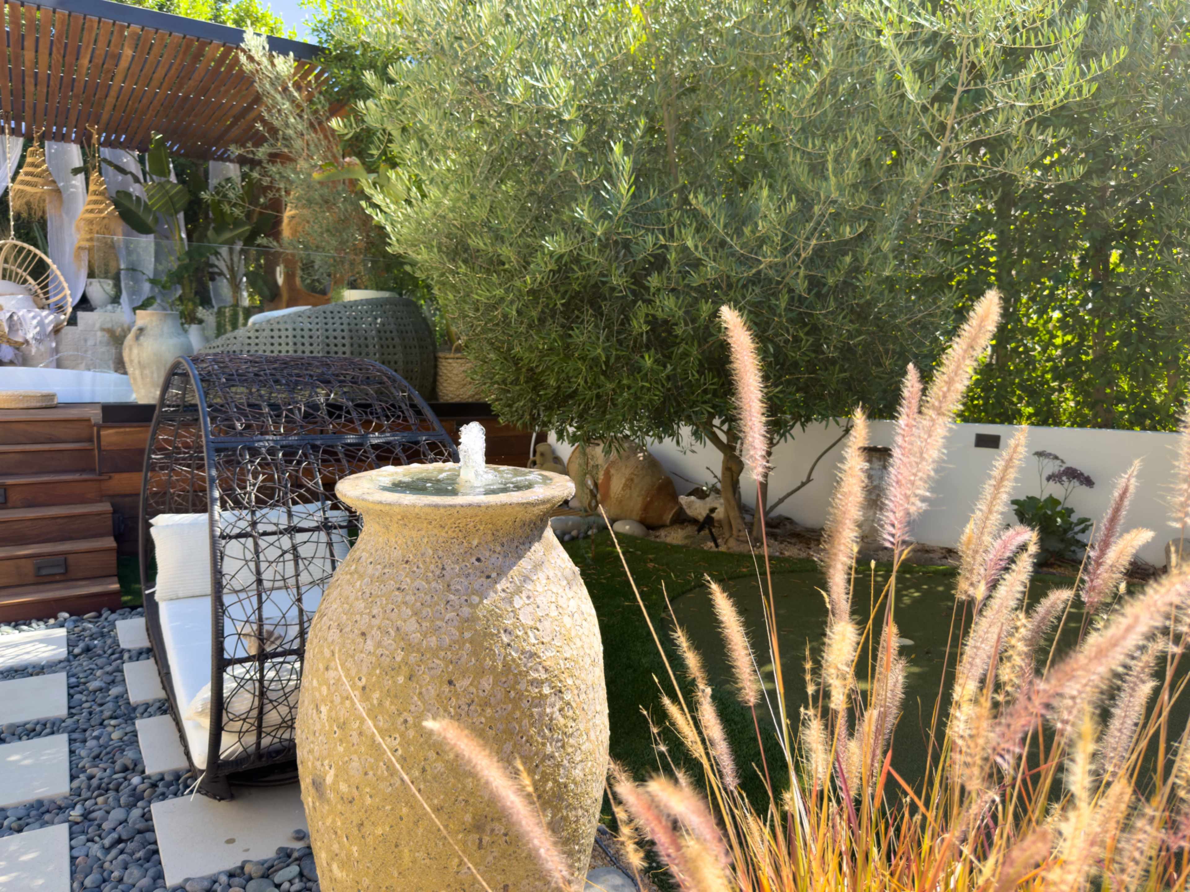 The image depicts a serene garden setting featuring a decorative stone fountain, a circular wicker chair, and lush greenery, including ornamental grasses and an olive tree.