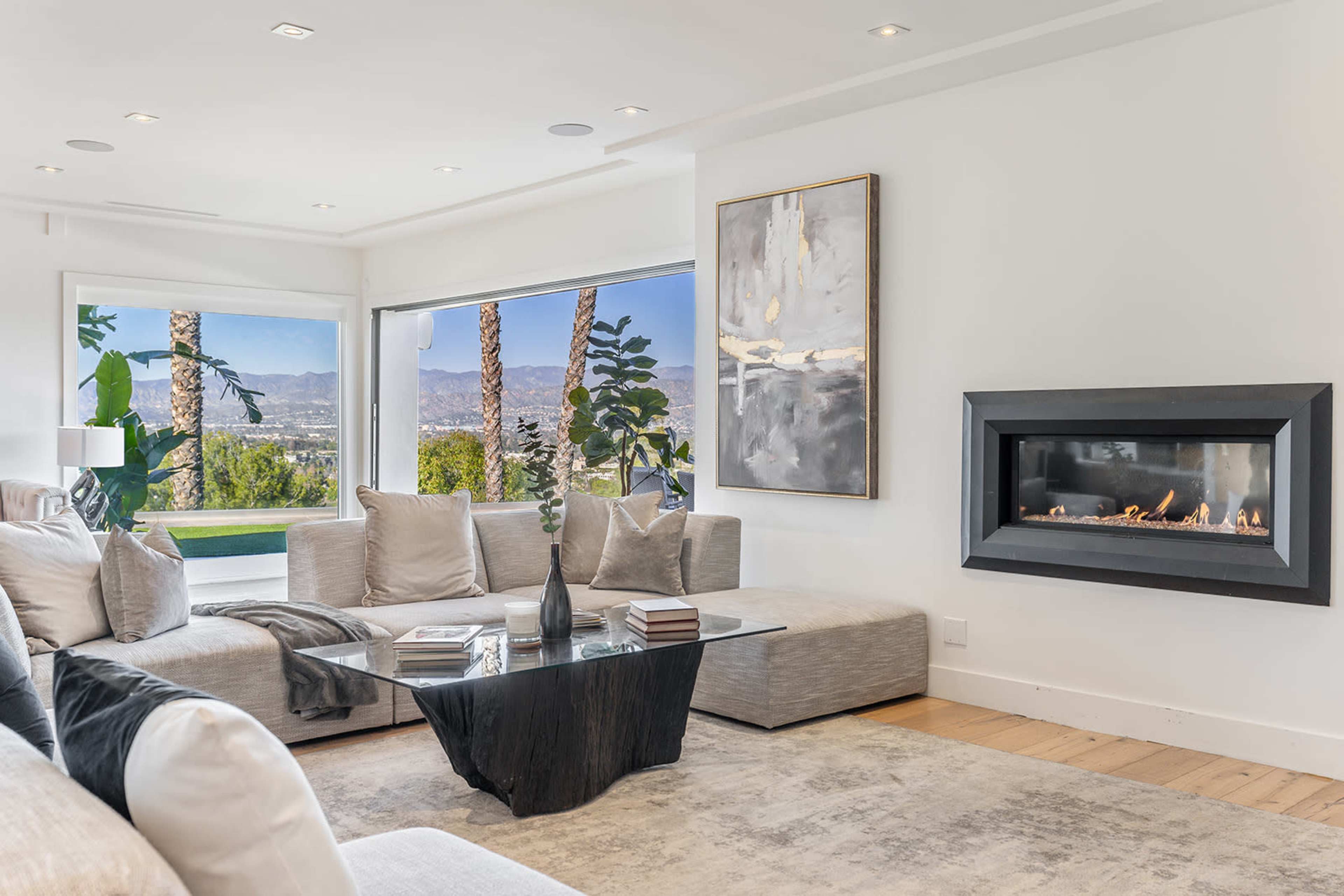A modern living room features a sectional sofa, a black coffee table, a fireplace, and large windows showcasing a scenic view of palm trees and mountains.