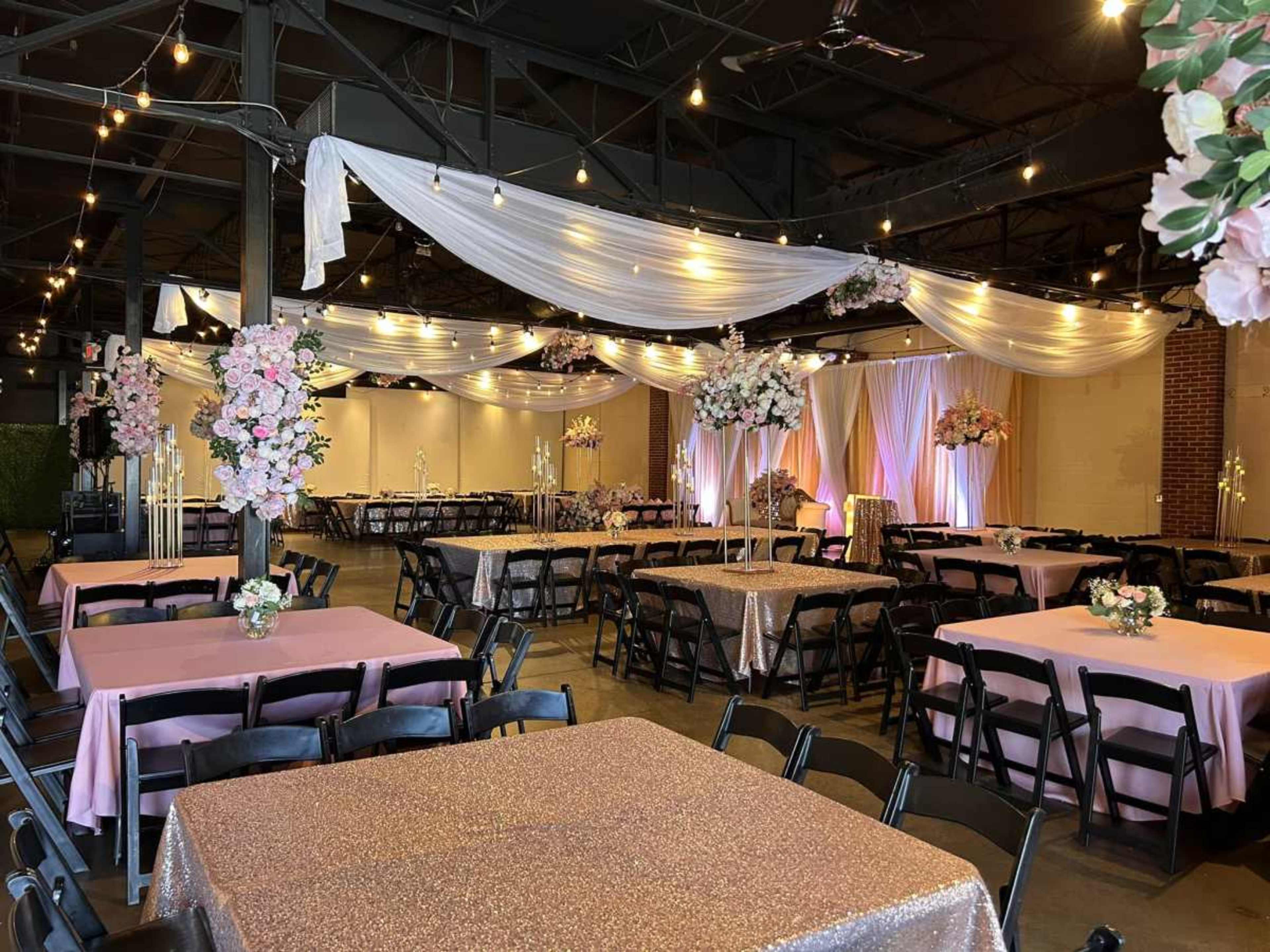 A spacious decorated event venue features elegantly arranged tables with pink tablecloths, floral centerpieces, and draped fabric overhead.
