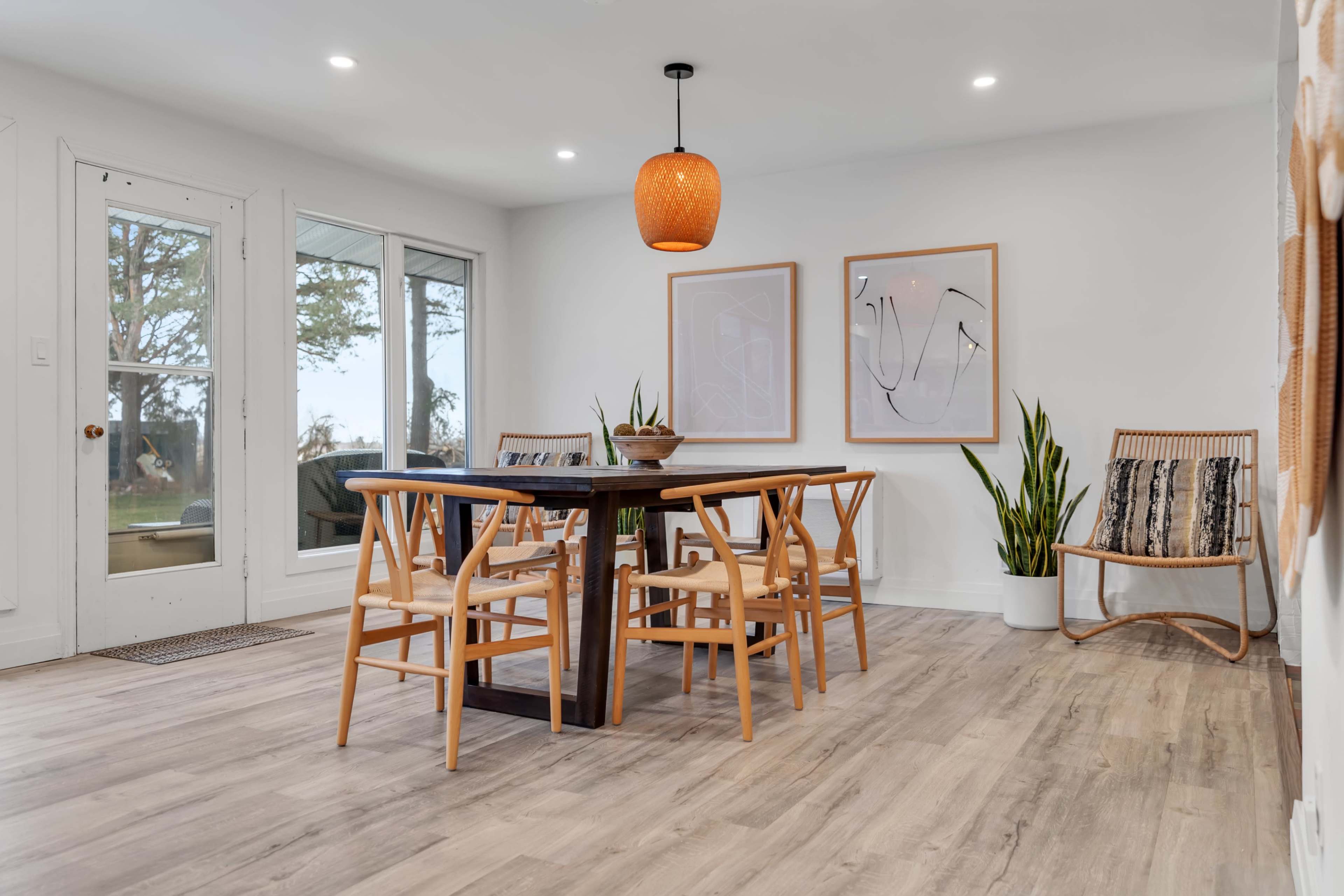 A dining area features a wooden table surrounded by chairs, with a pendant lamp overhead and plants positioned in the corners.