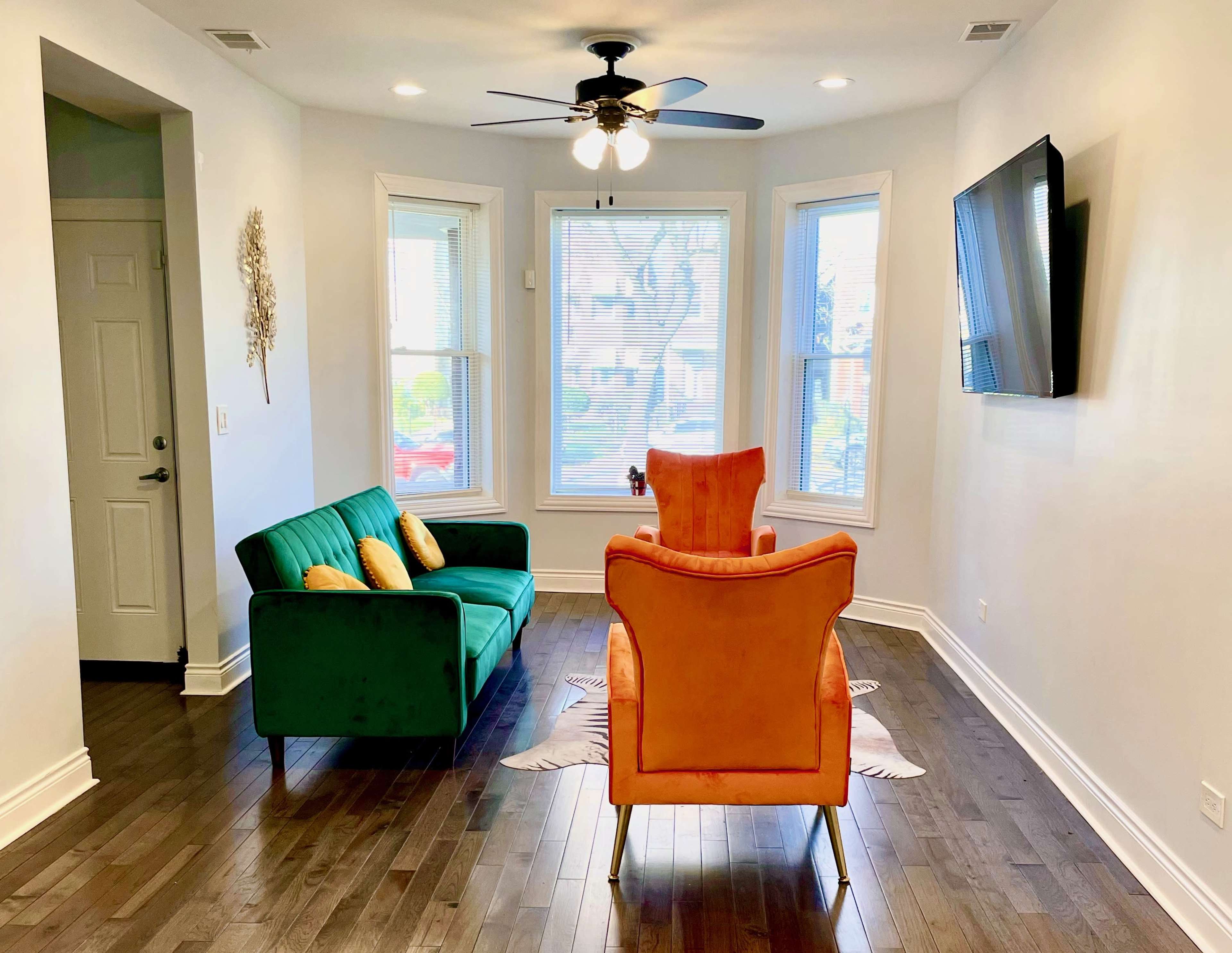 A living room with a green couch, an orange armchair, hardwood floors, large windows, and a mounted television on the wall.