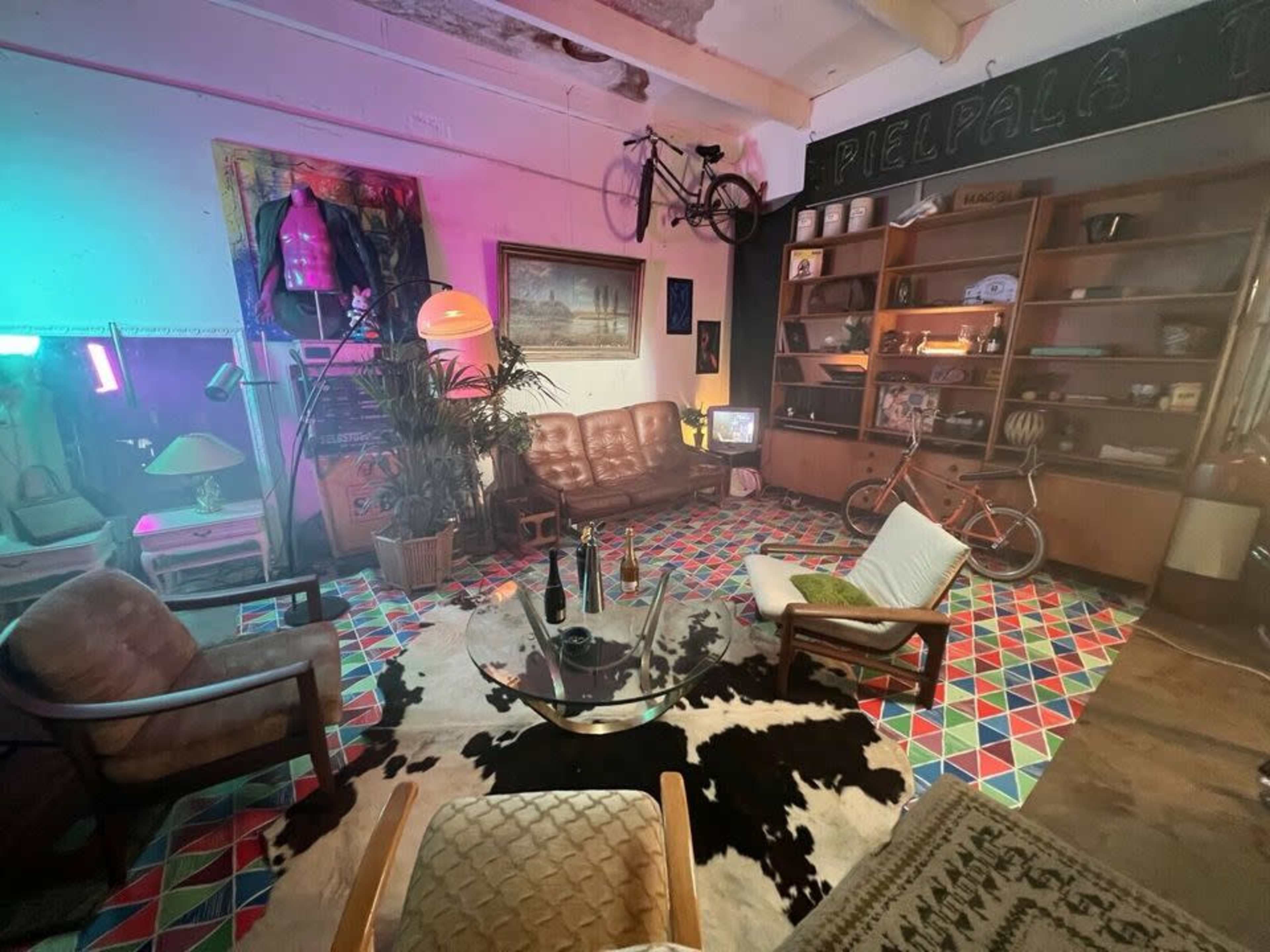 A retro-styled indoor living space with patterned flooring, vintage furniture, and a bicycle hanging from the ceiling, illuminated by colorful lights.