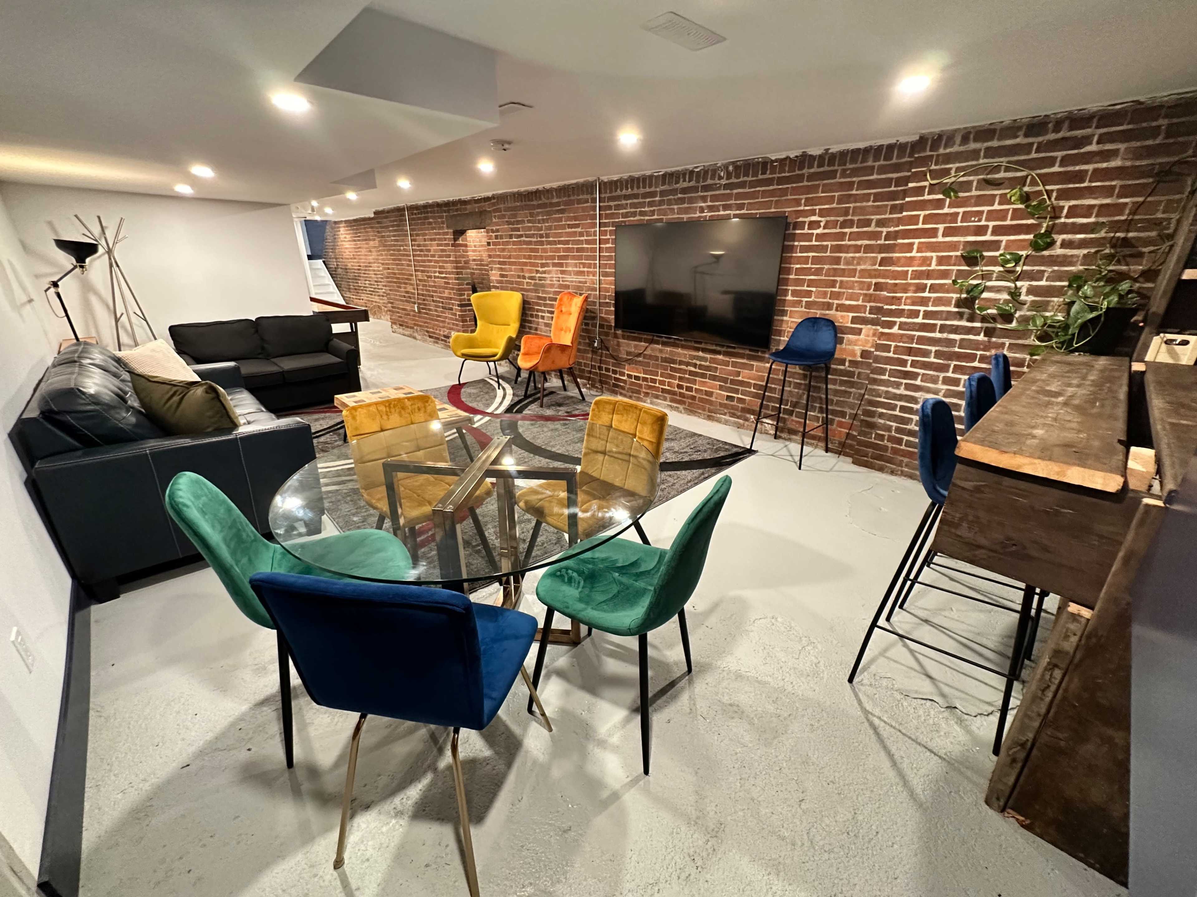 A modern basement living area with a seating arrangement that includes a black sofa, colorful chairs, a glass dining table, and a wall-mounted television.