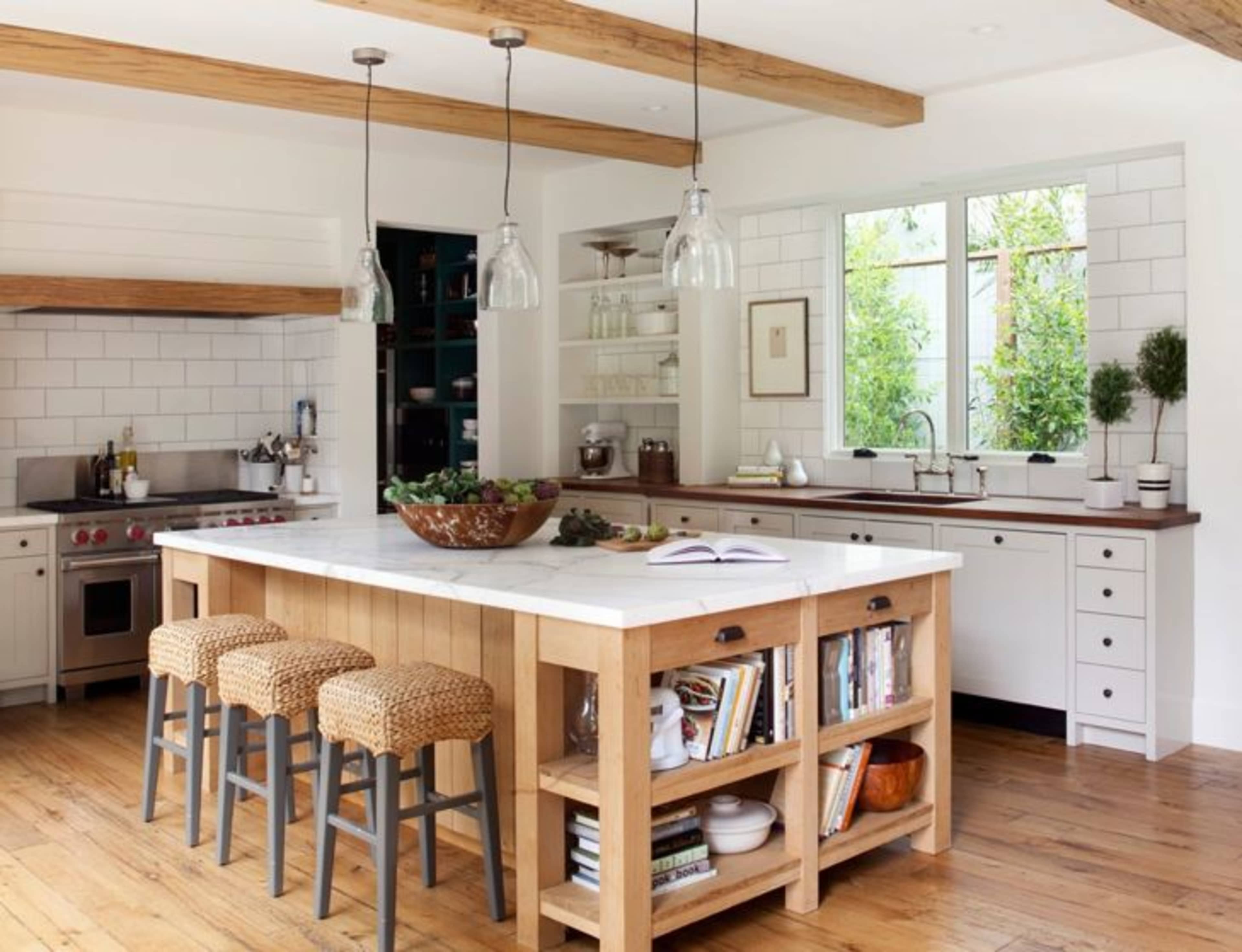 The kitchen features a large central island with a marble countertop, surrounded by woven bar stools, and is complemented by wooden beams and a window overlooking greenery.