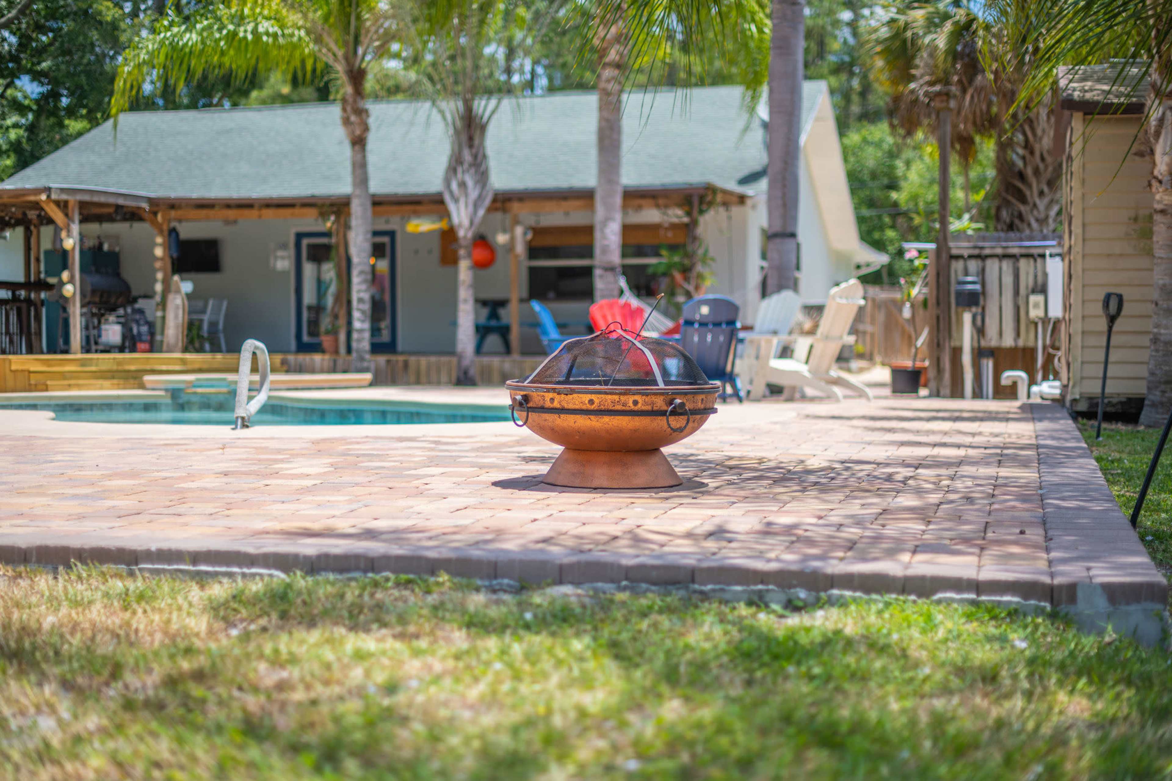A decorative fire pit sits on a paved patio surrounded by palm trees and a swimming pool, with a house visible in the background.