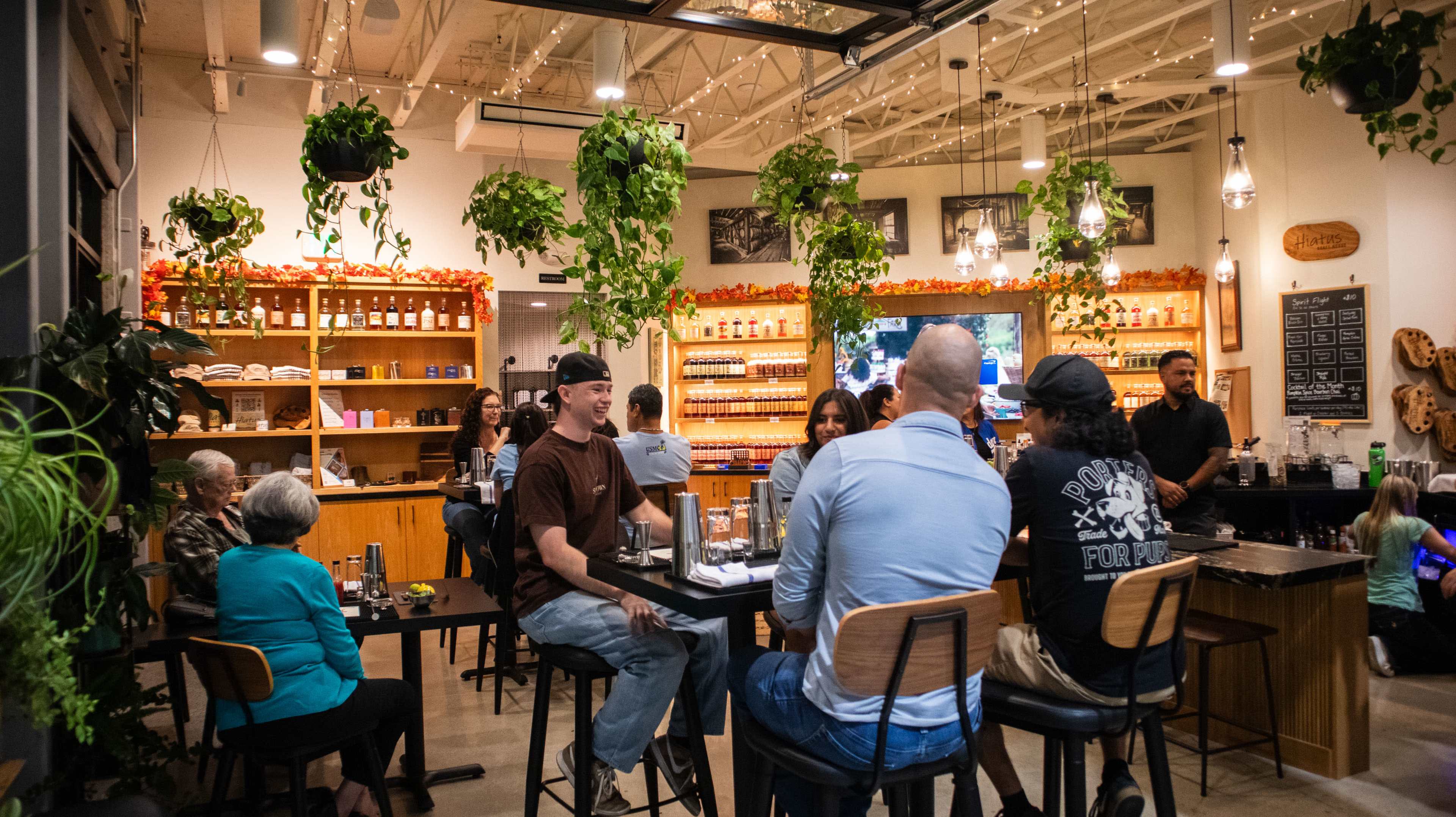 A group of people are seated around a bar in a vibrant indoor space filled with plants and wooden shelves stocked with various bottles.