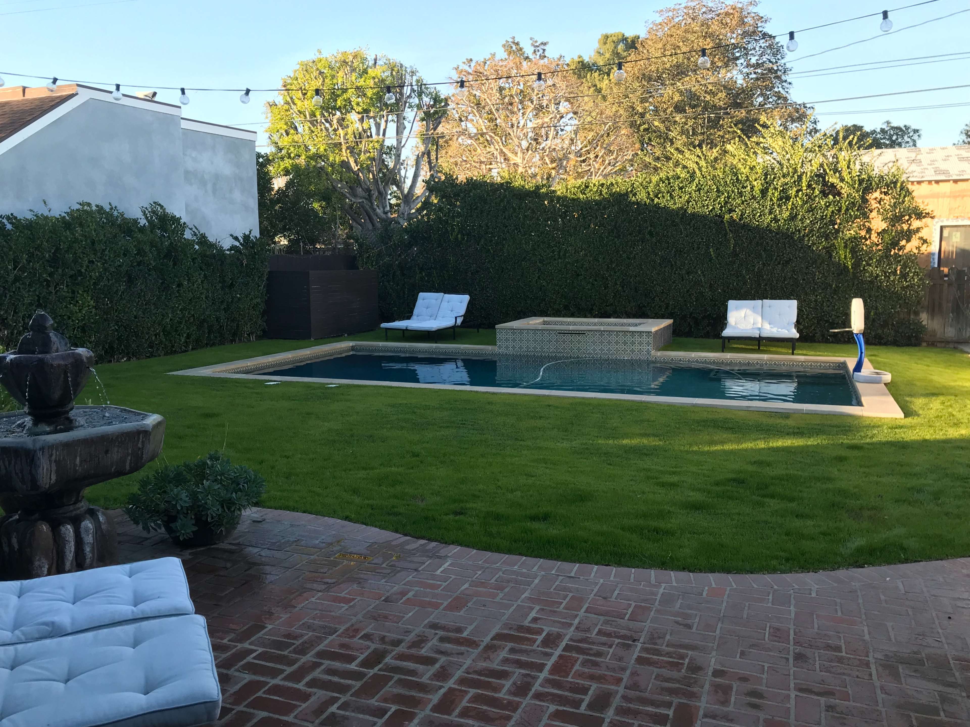 The image shows a backyard with a swimming pool surrounded by lounge chairs, a fountain, and neatly trimmed grass.