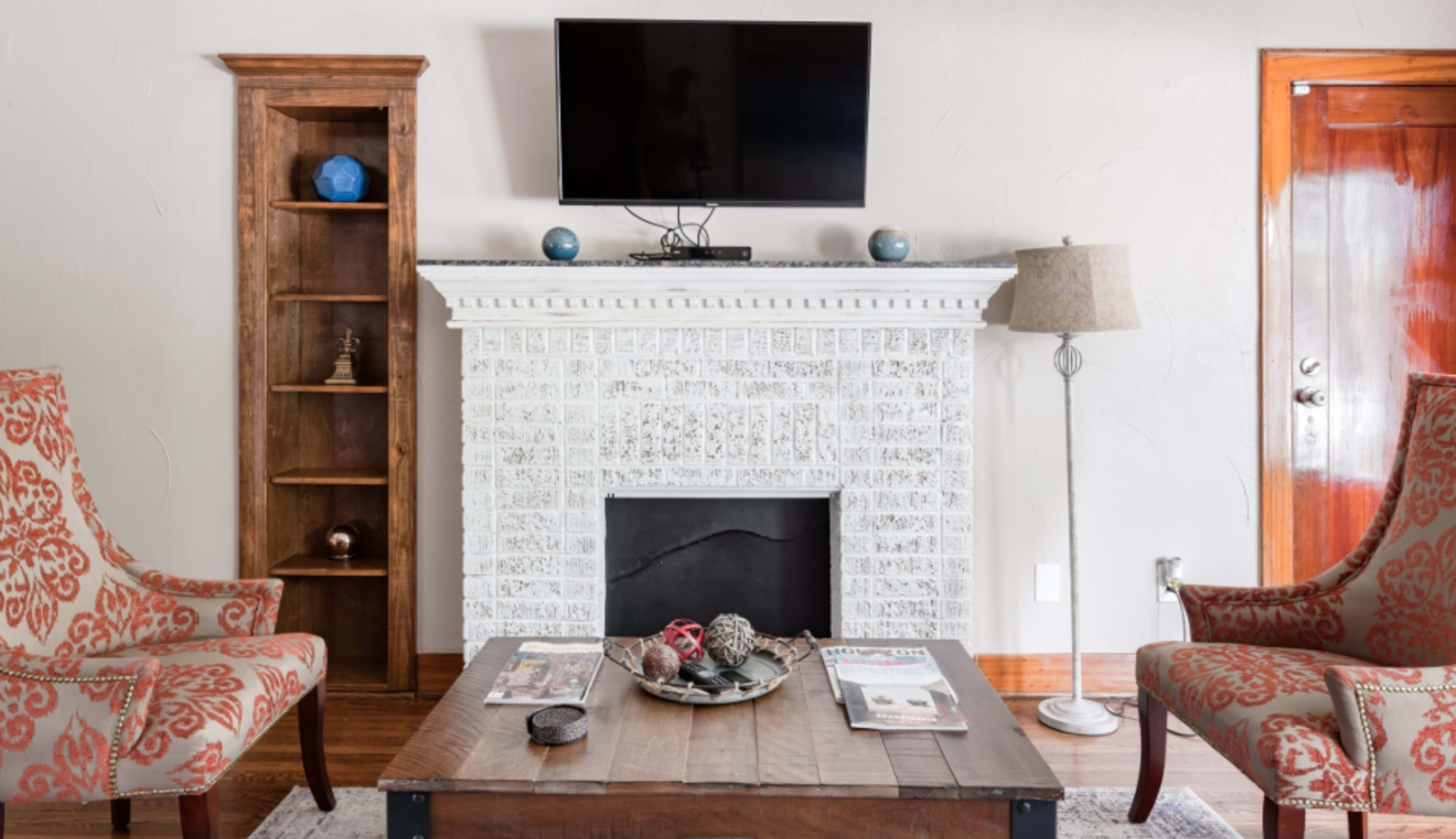 A living room with a fireplace, a mounted television above it, two patterned armchairs, and a wooden coffee table with magazines and decorative items.