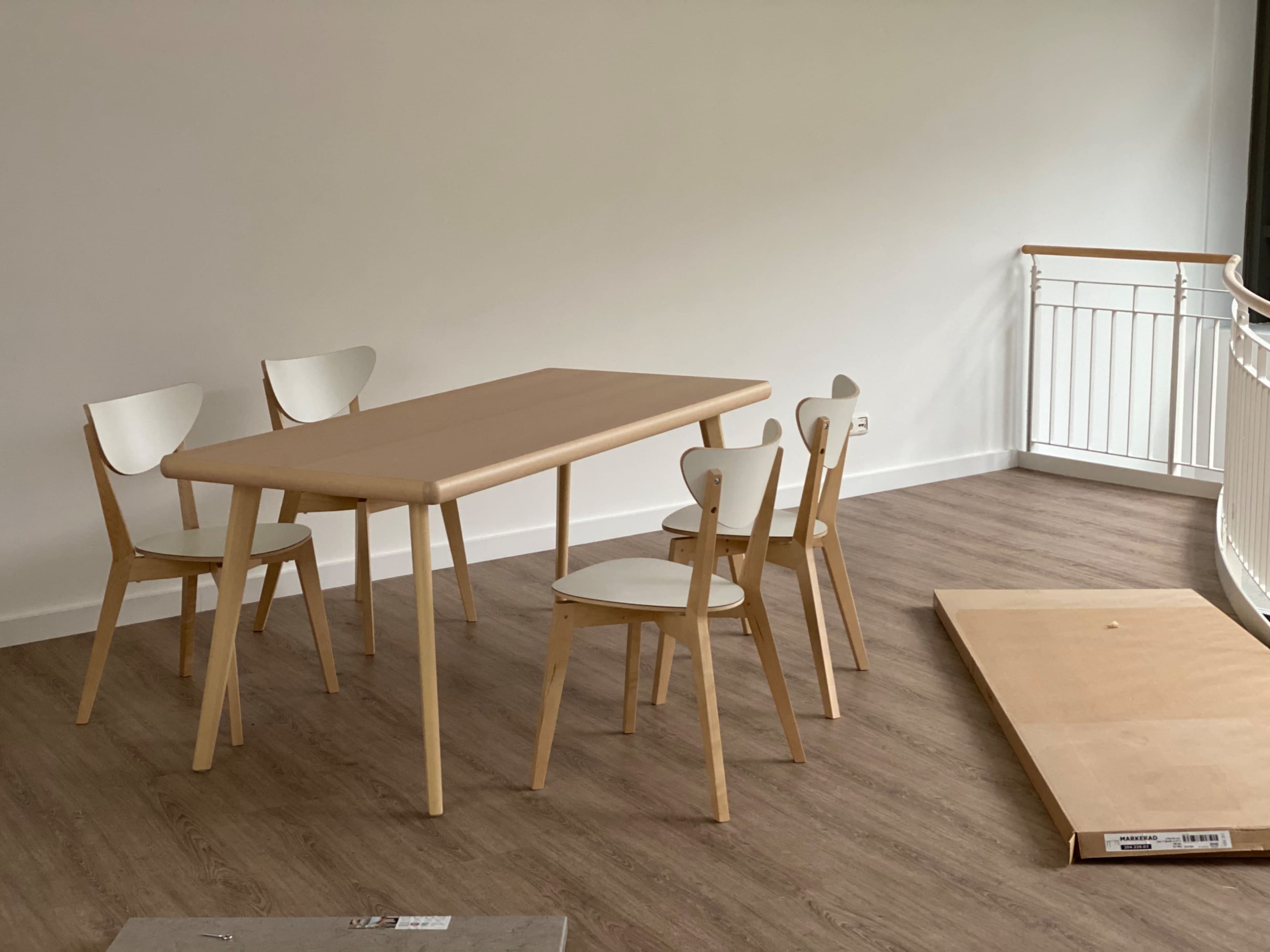 A wooden dining table is set with four matching chairs in a brightly lit room with light-colored flooring and a railing.