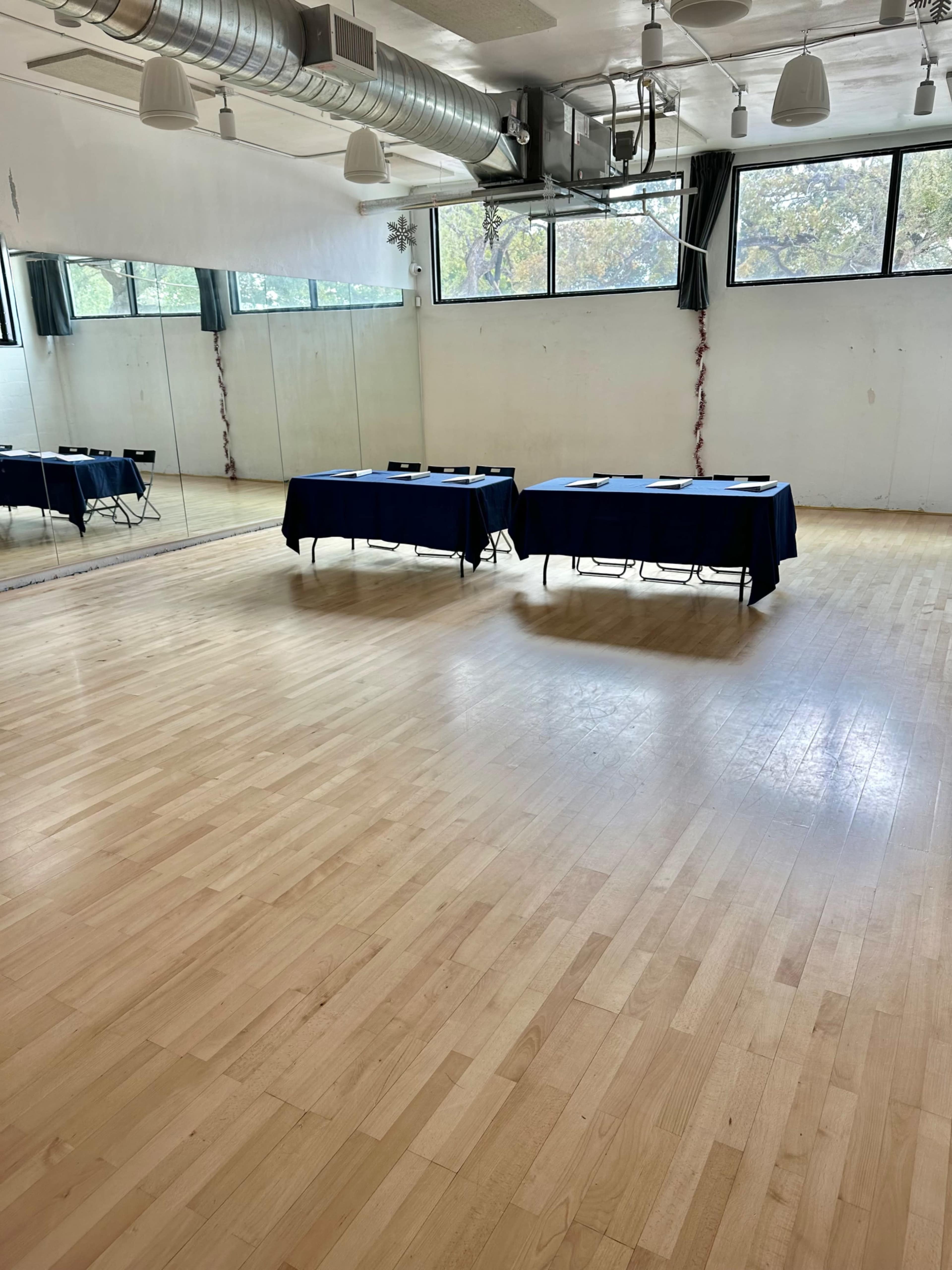 The image shows a spacious room with wooden flooring, featuring two tables covered with blue cloths and several chairs arranged around them, along with large windows allowing natural light.