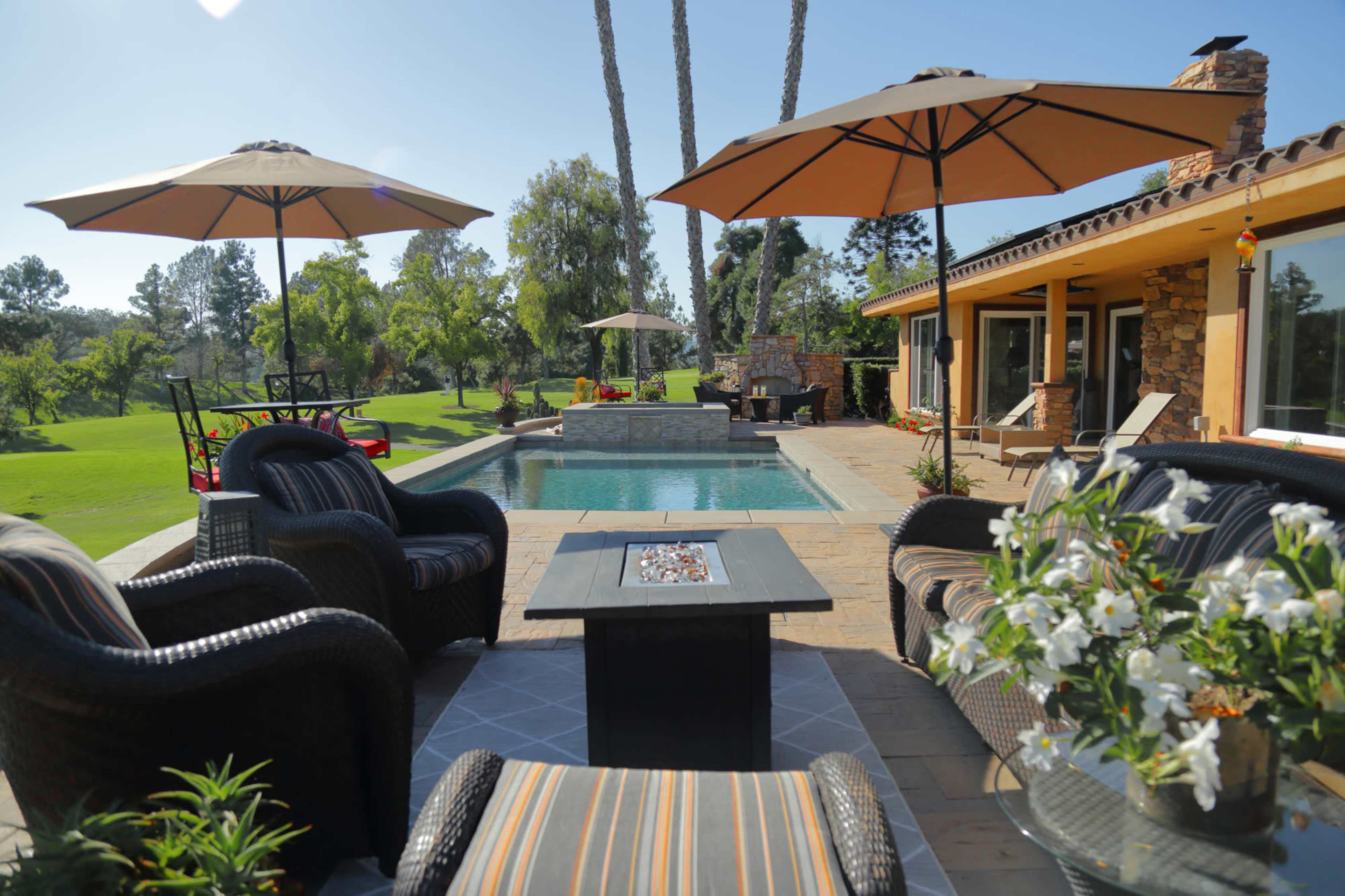 A landscaped outdoor area with a swimming pool surrounded by lounge chairs and umbrellas, alongside a view of a manicured lawn and trees.
