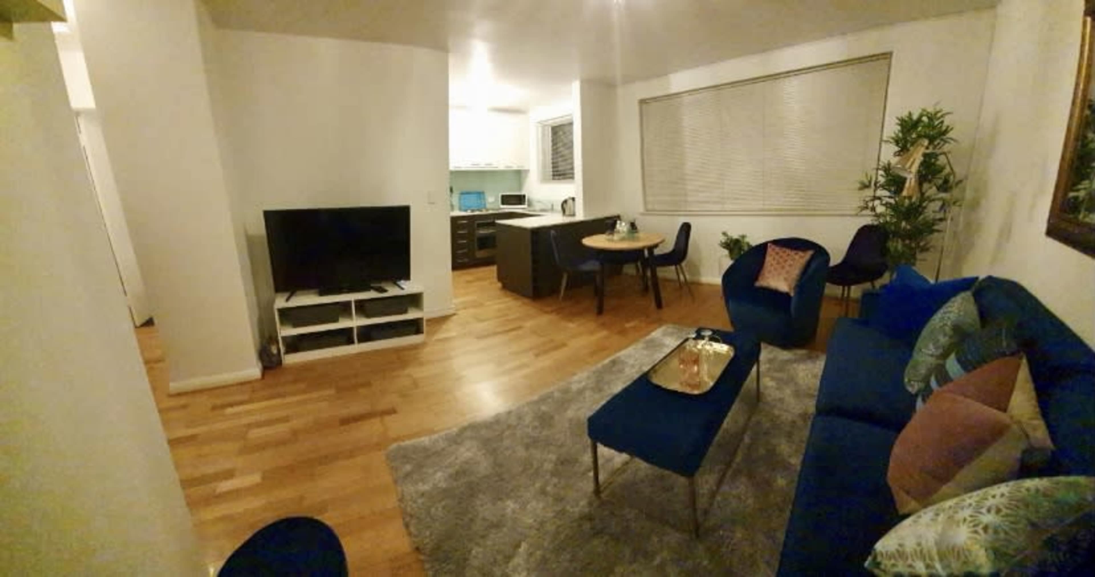 A modern living space featuring a seating area with blue chairs and a sofa, a television on a stand, and a dining table in an open kitchen setting.