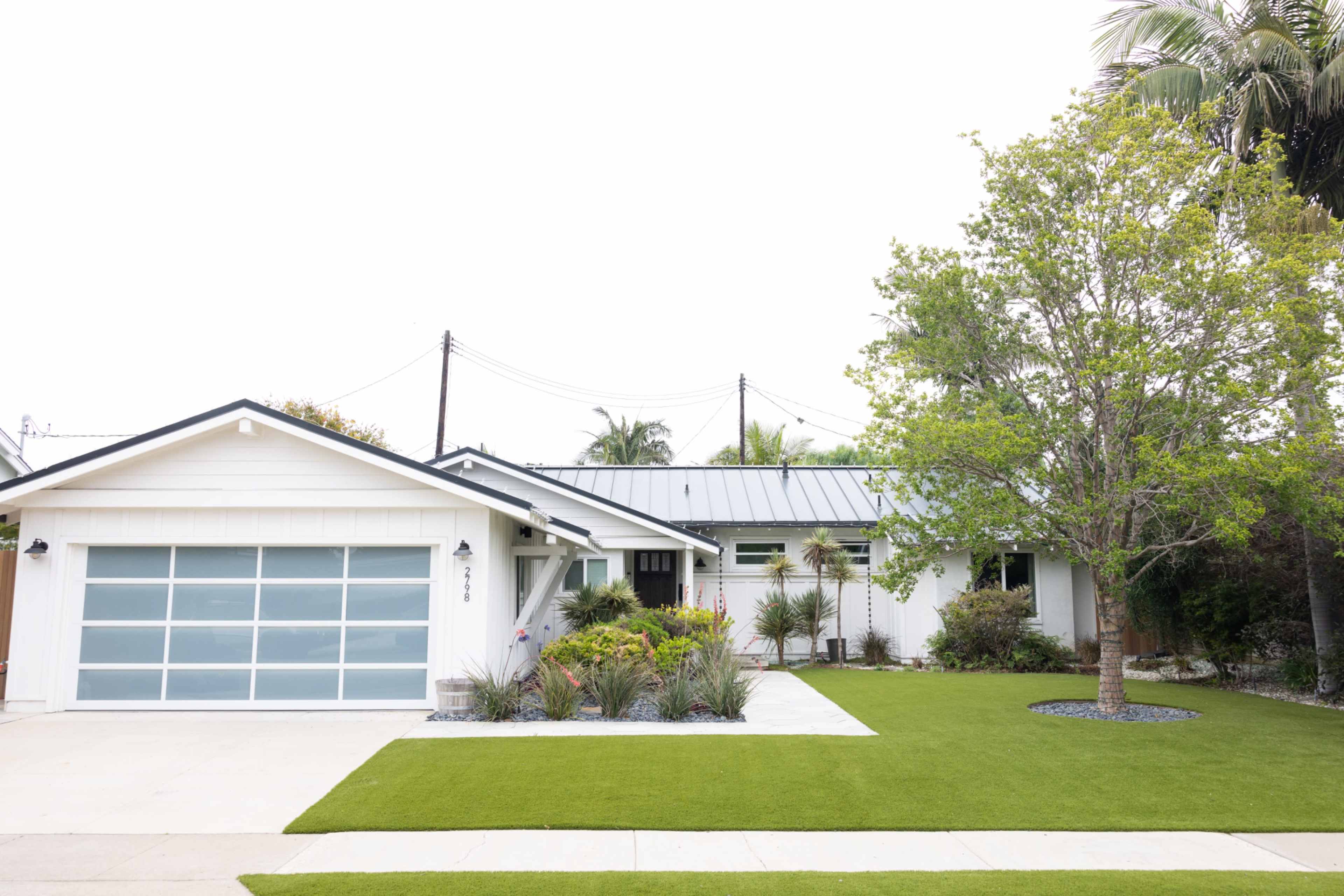 A white single-story house with a gray garage door, surrounded by green landscaping and a palm tree in the front yard.