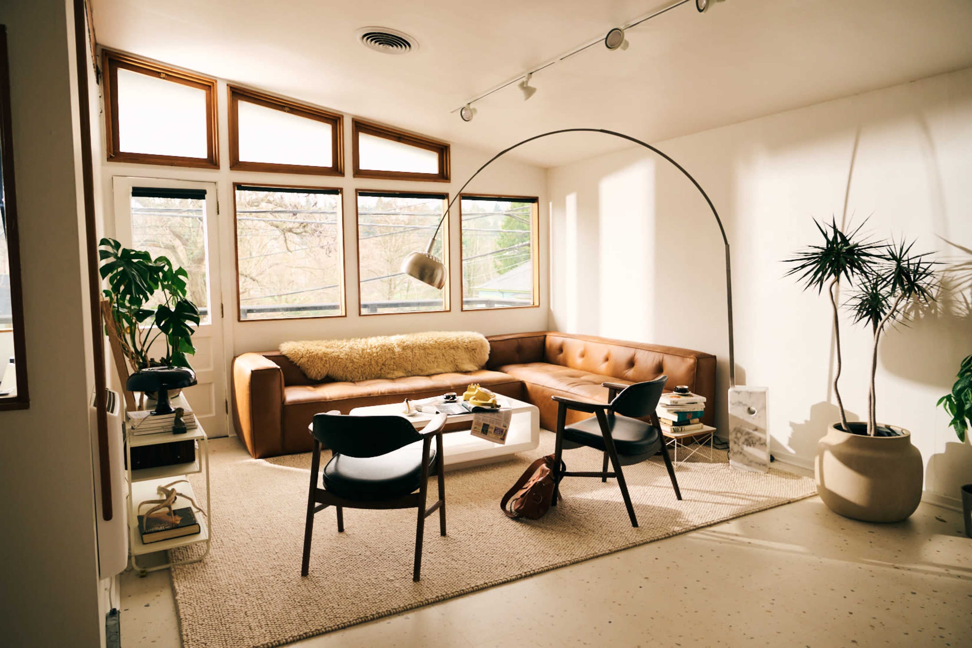 The image shows a modern living room featuring a brown leather sofa, a black chair, and large windows that allow natural light to illuminate the space.