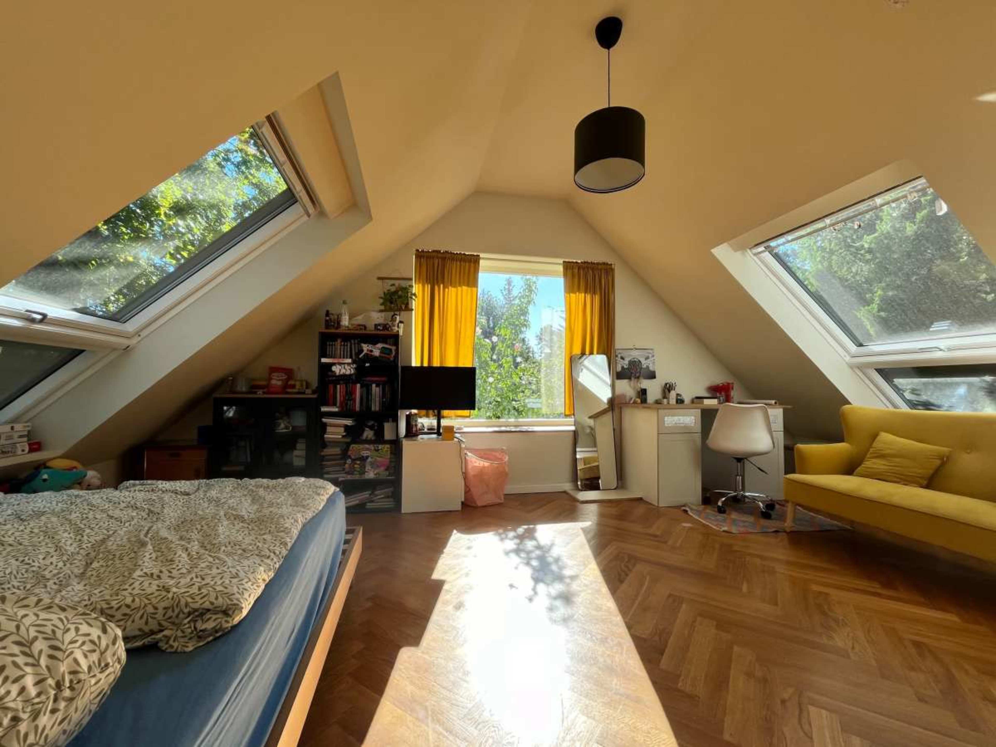 A bright, slanted-roof bedroom features a bed, a desk with a chair, and a yellow couch, illuminated by natural light streaming through large windows.