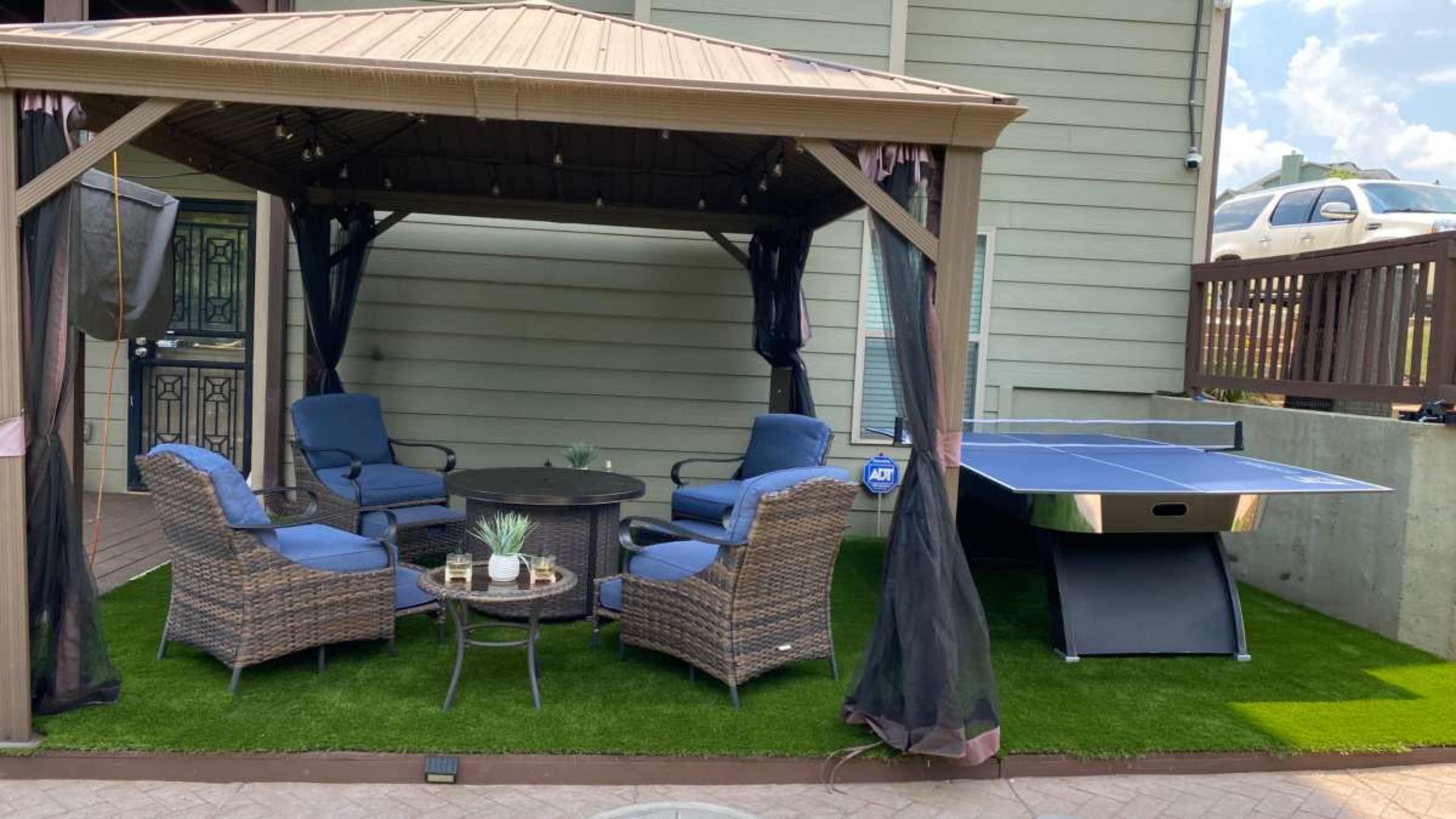 A gazebo with four blue chairs and a table, adjacent to a blue ping pong table on an artificial grass surface.