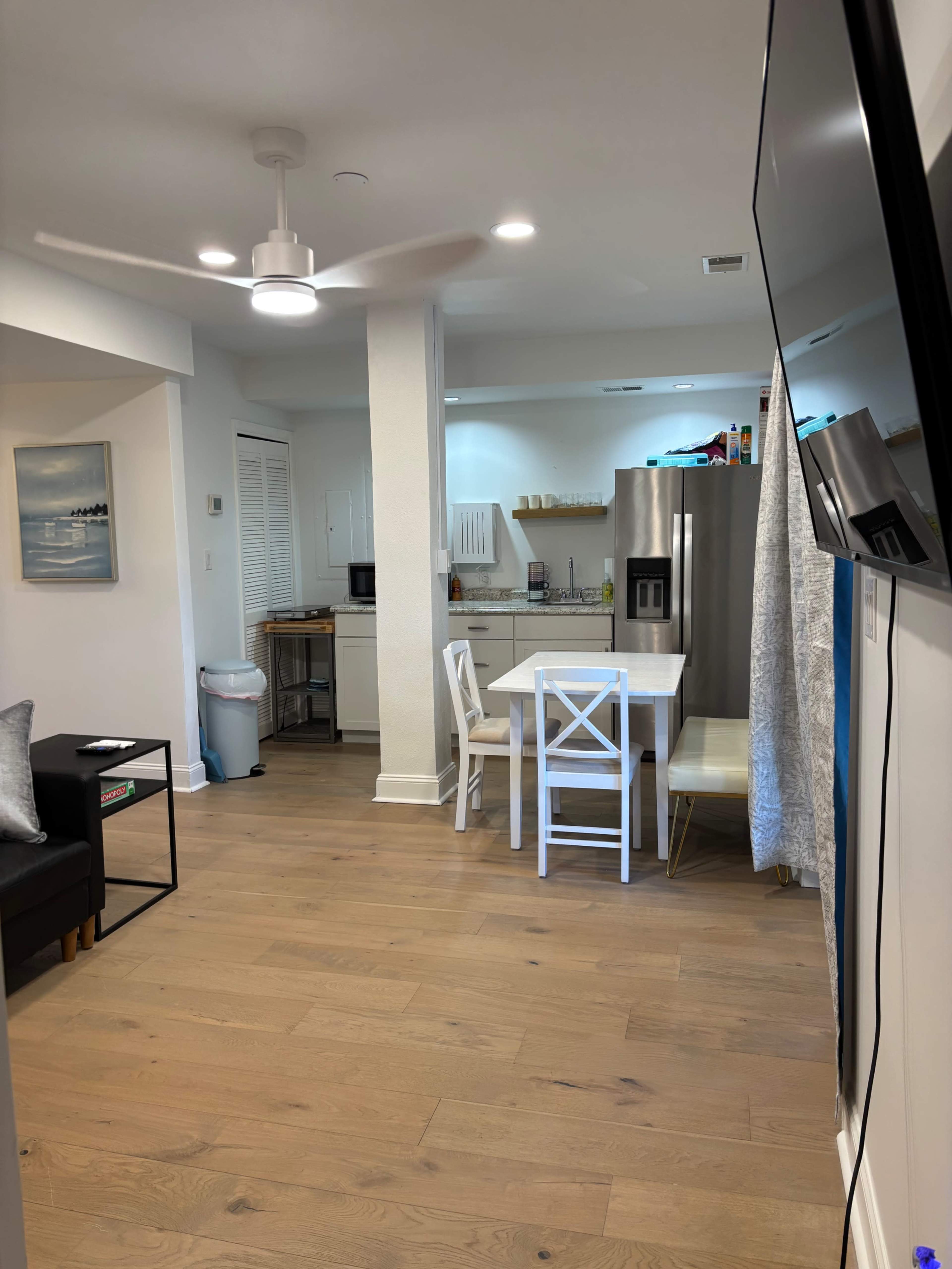 The image shows a modern, open-plan living area leading to a kitchen with stainless steel appliances and a dining table set for four.